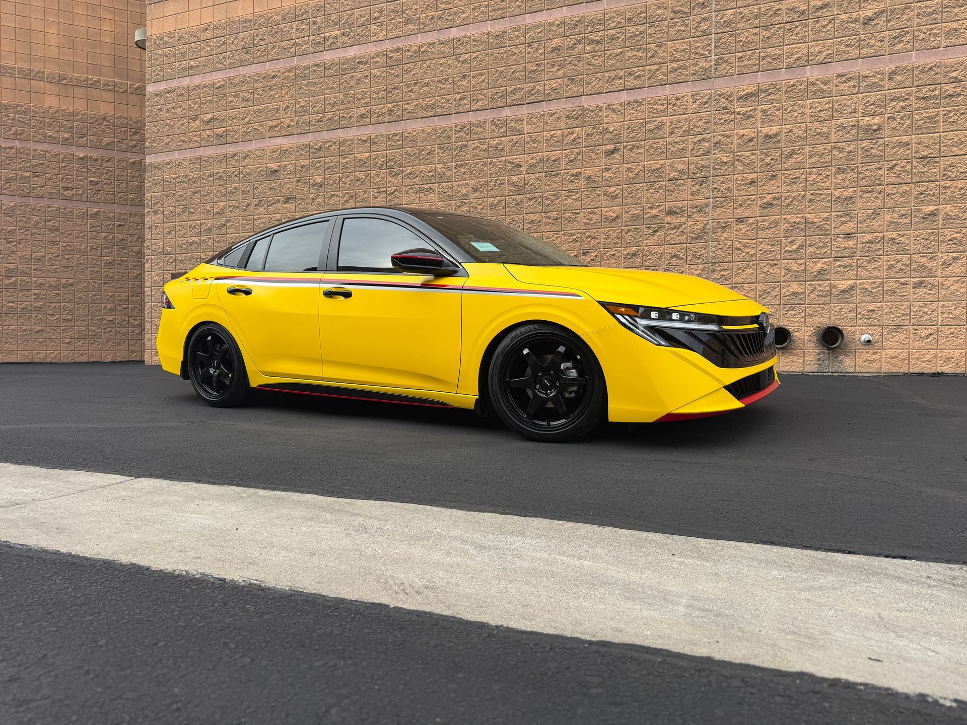 Yellow sports car parked beside a tan brick wall on dark asphalt, with black wheels and a black roof.