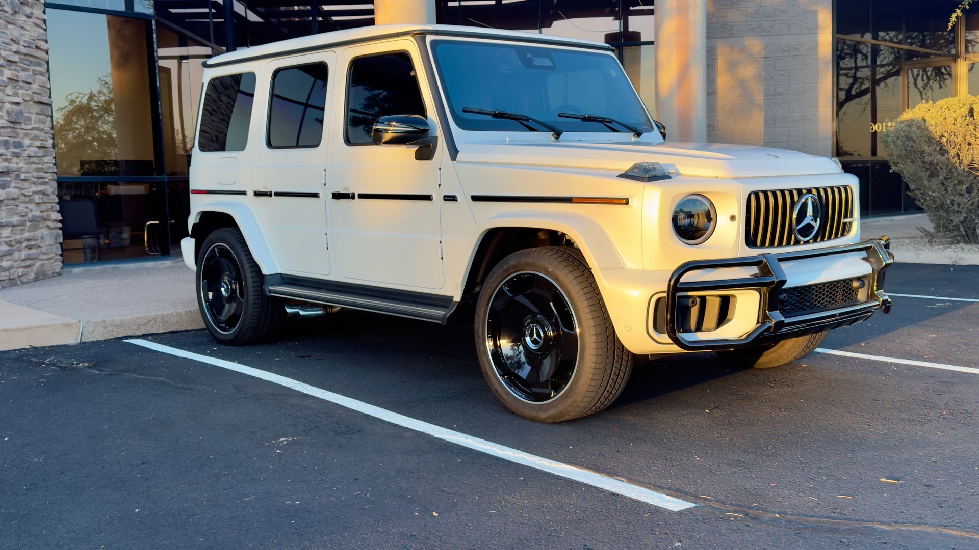 White Mercedes G-Class SUV parked in a lot beside a building, viewed from the front-left side.