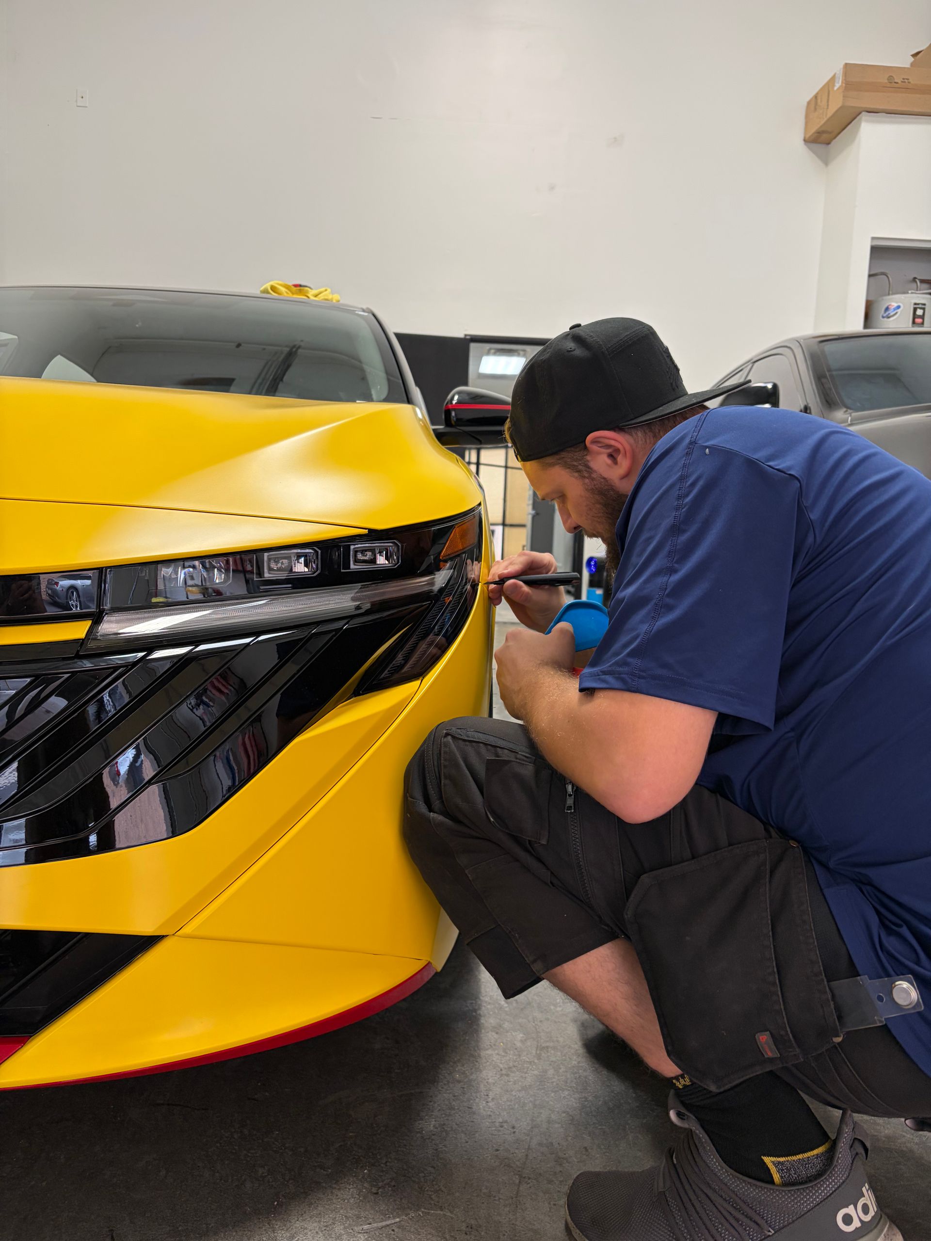 Technician crouches beside a yellow sports car, inspecting or repairing the front bumper in a garage.