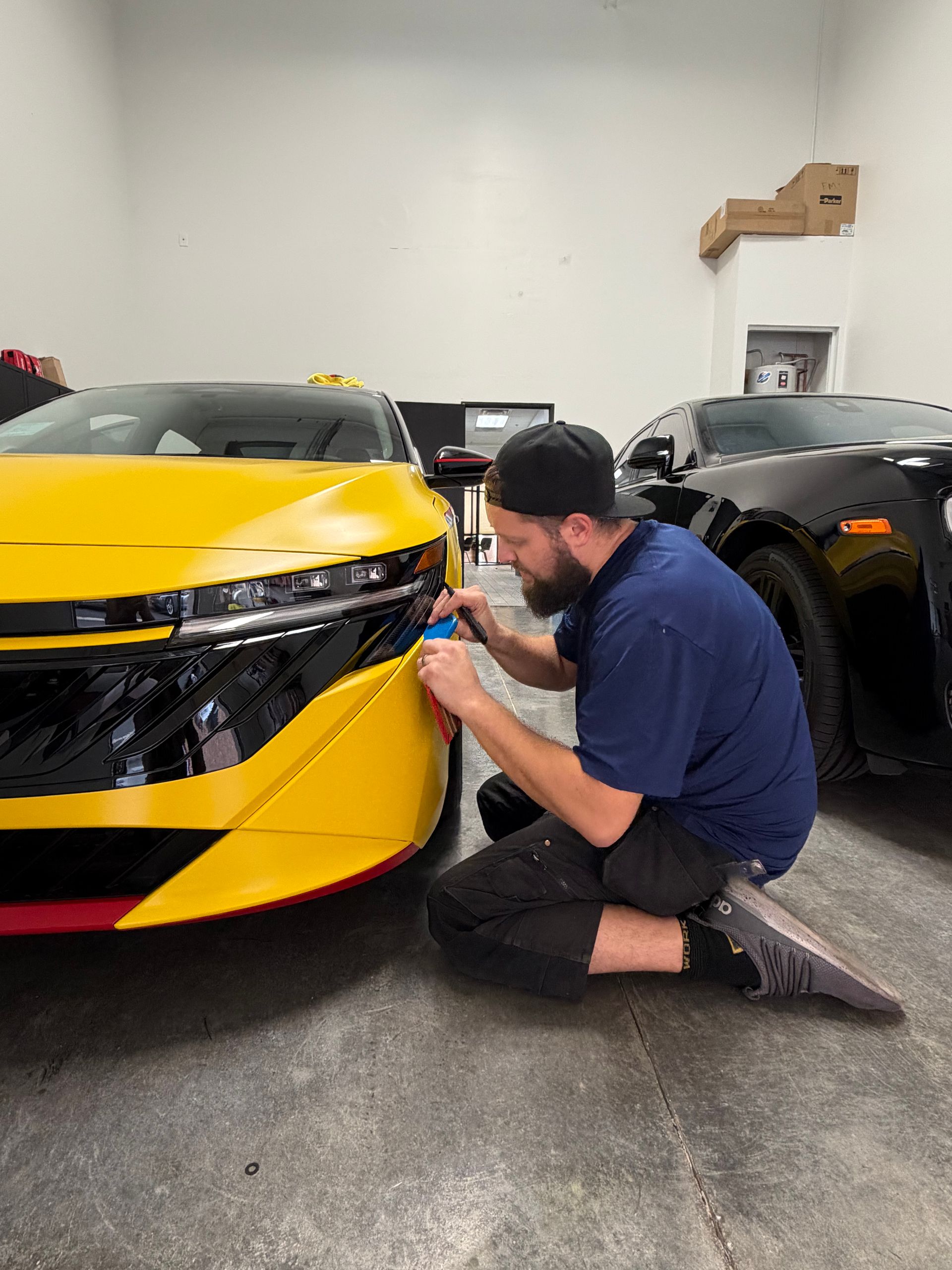 Mechanic kneeling beside a yellow sports car, adjusting the front bumper in a garage.