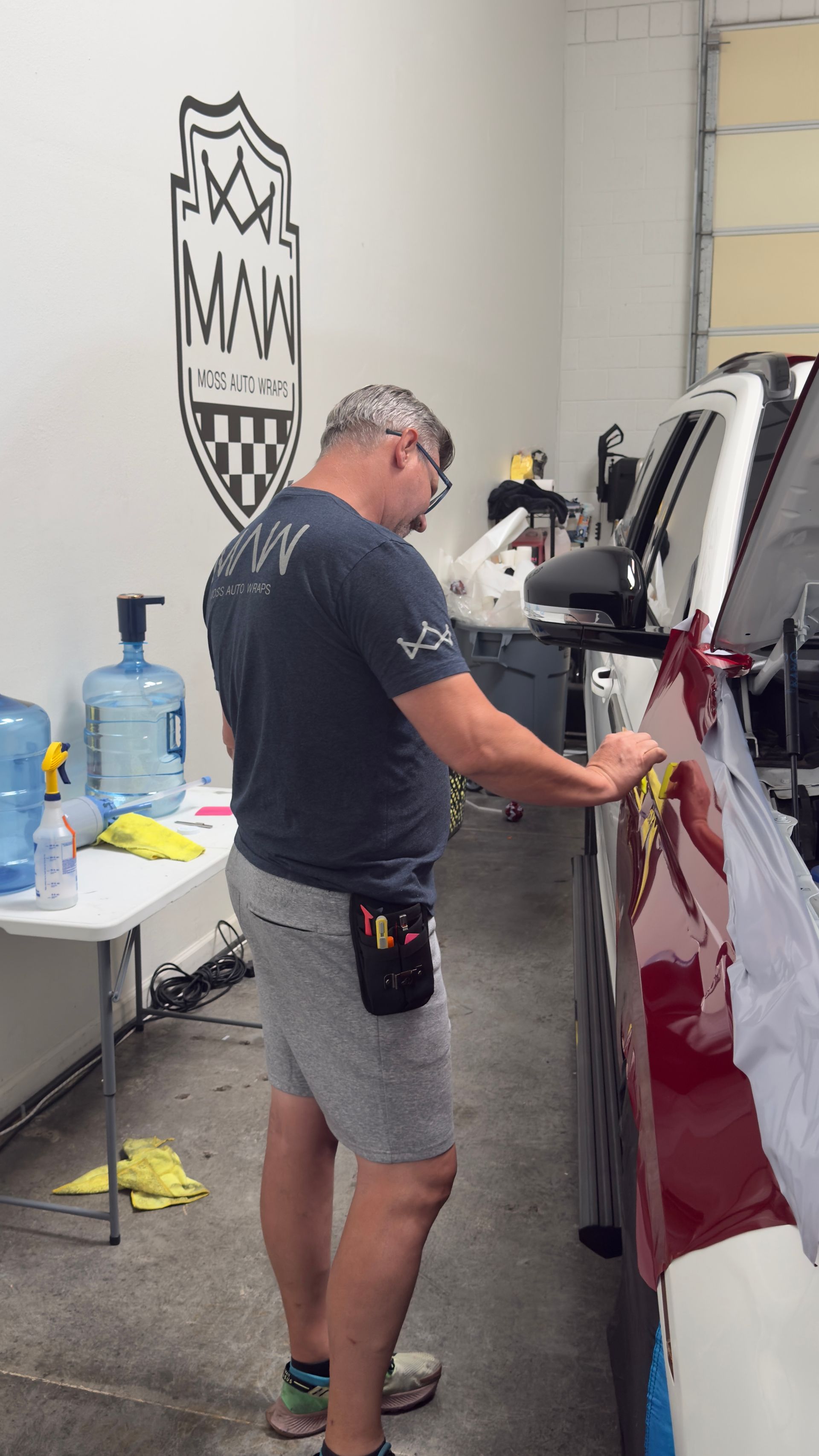 Person working on a maroon car in an auto repair shop, polishing the side panel.