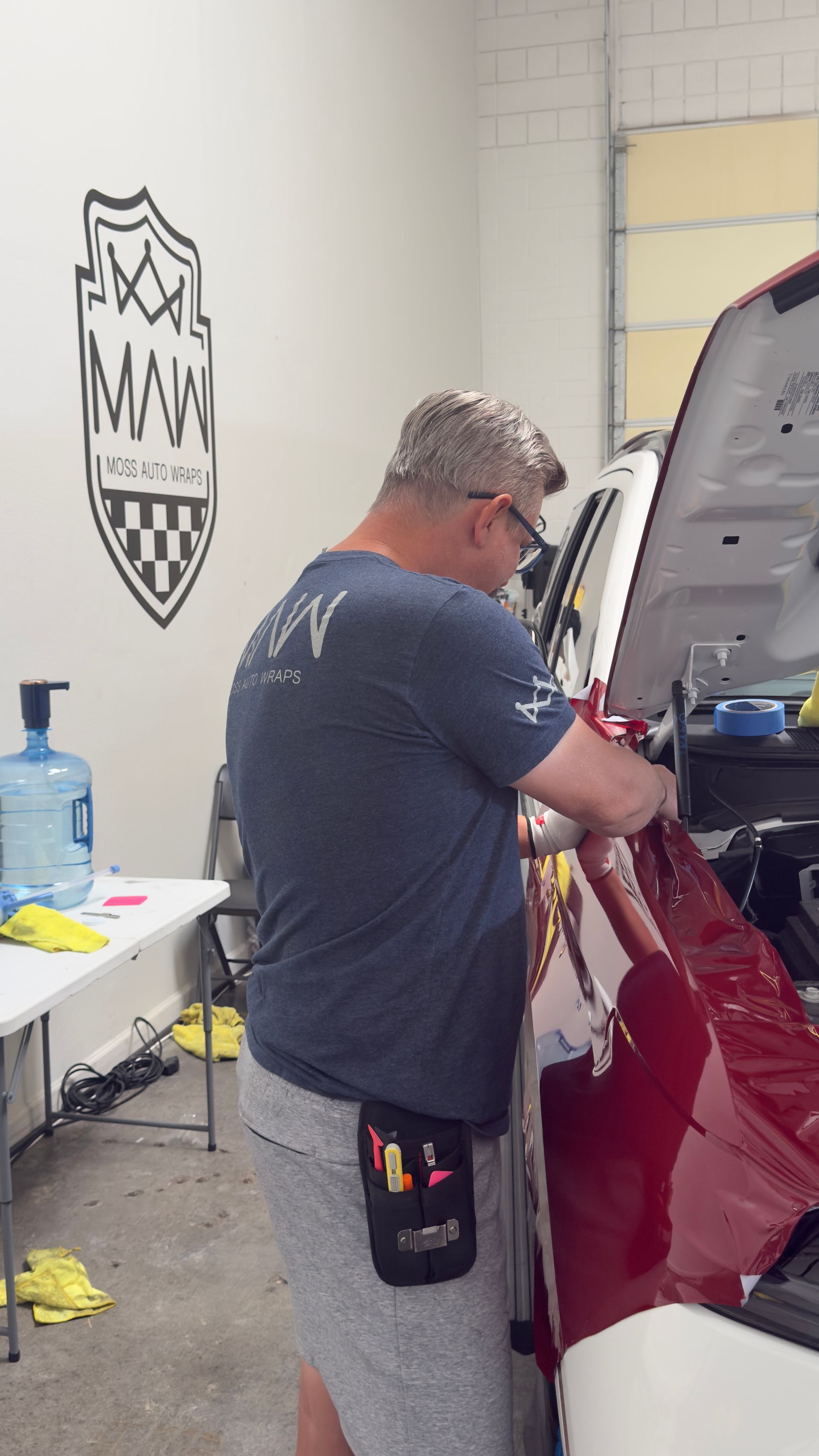 Man working on a red car in a garage beside a table with supplies and a blue water jug