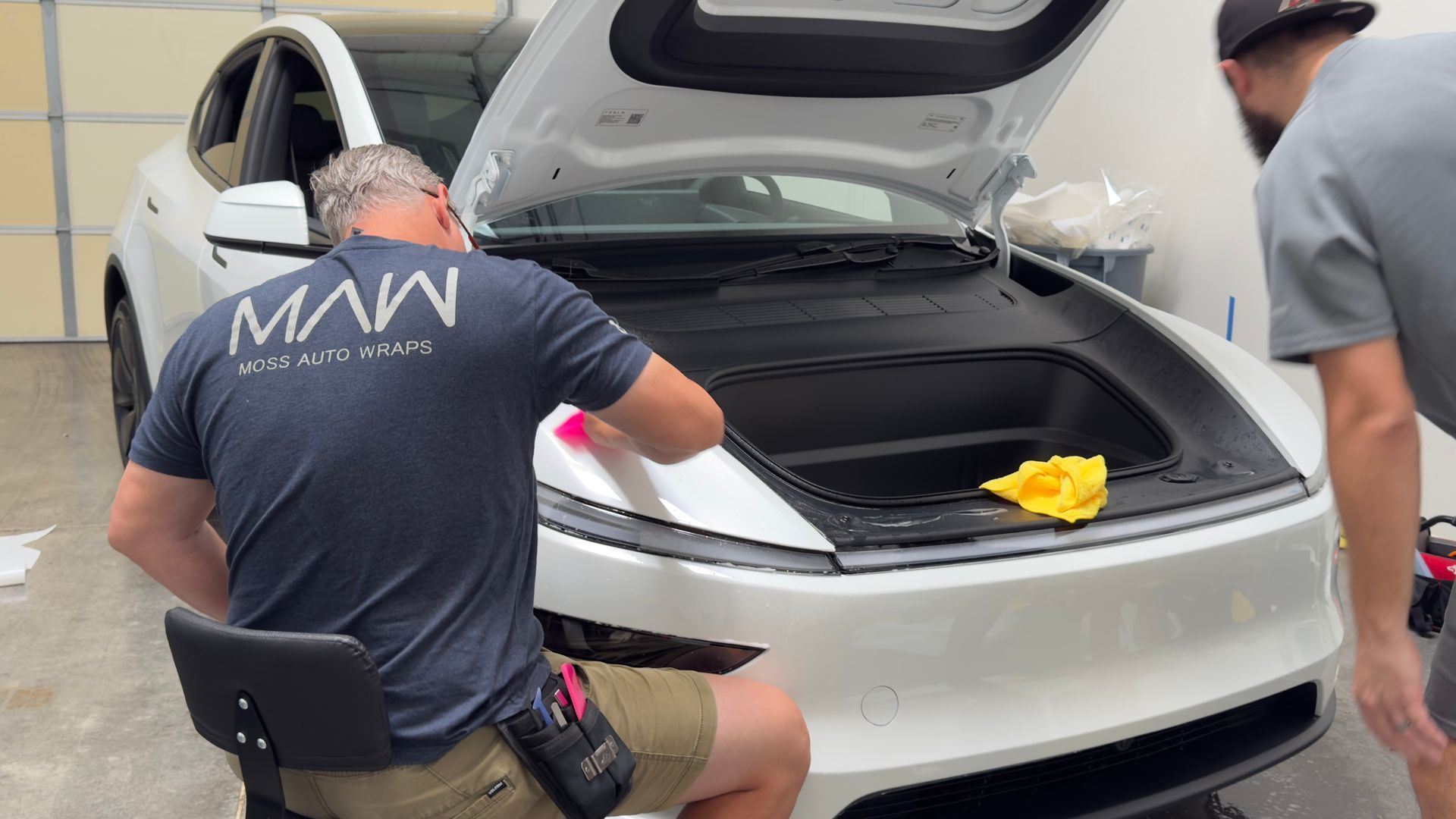 Two people cleaning a white car’s hood in a garage, with a yellow cloth on the front.