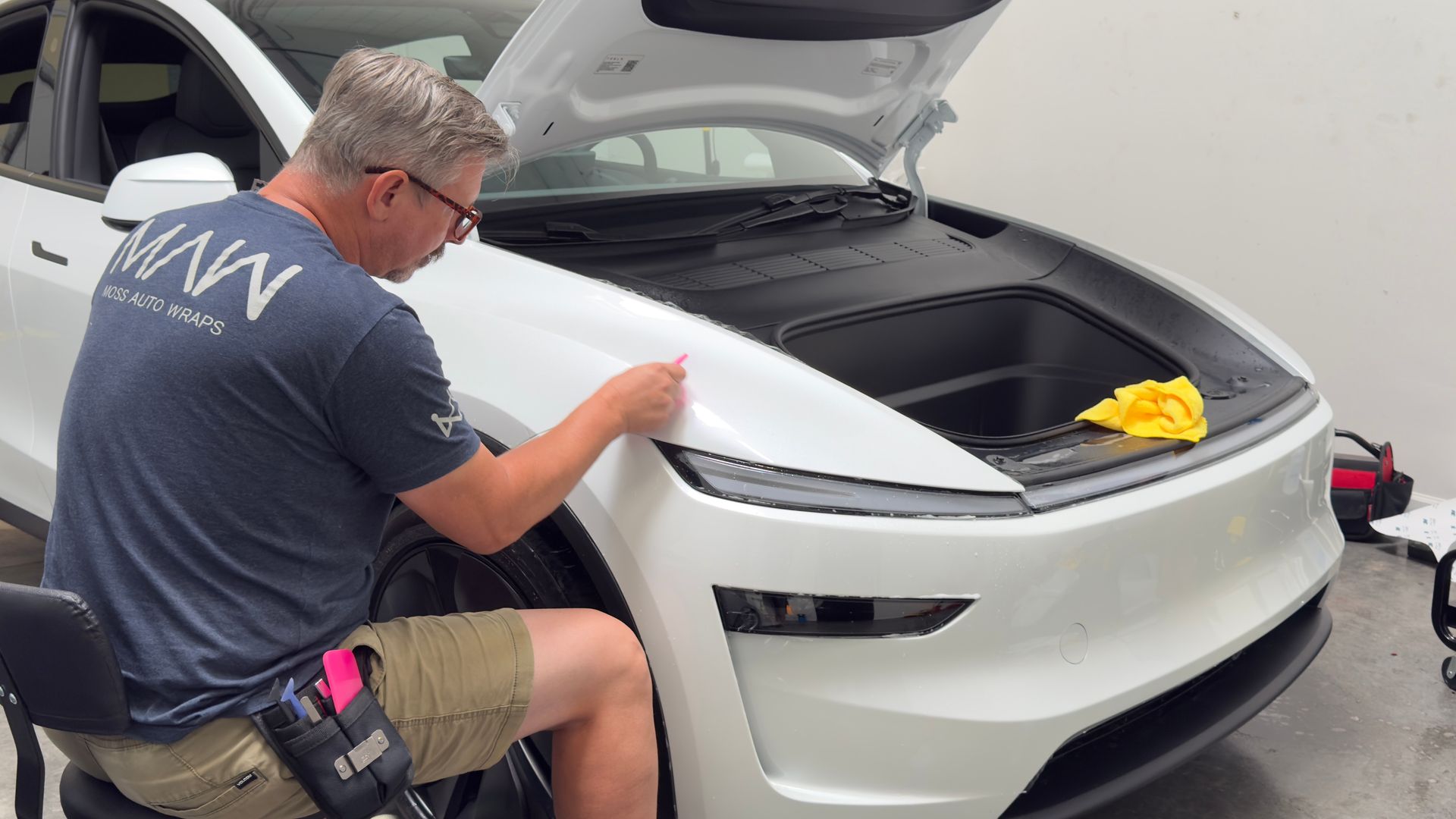 Man polishing the front hood of a white car in a workshop, with a yellow cloth nearby