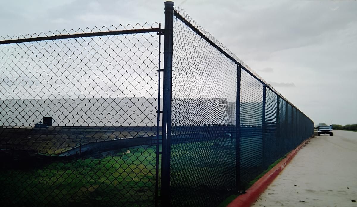 A long black chain-link fence with barbed wire along the top, bordering a grassy area and a road under a cloudy sky.