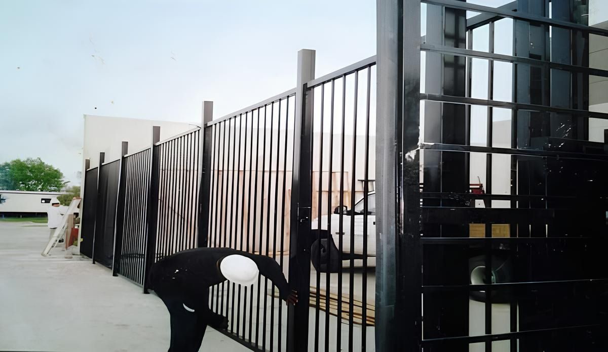 Person in dark clothing inspecting a black metal fence outdoors. The fence has vertical bars and a square post.