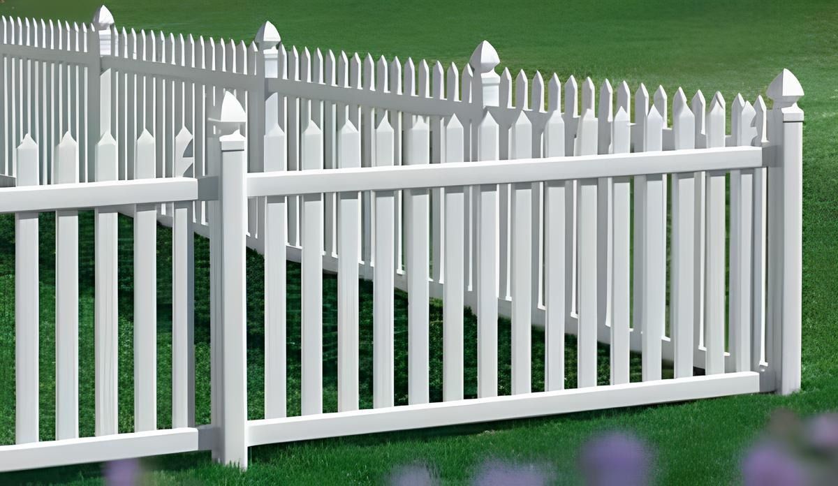 White picket fence surrounding a green lawn, with a few purple flowers visible in the foreground.