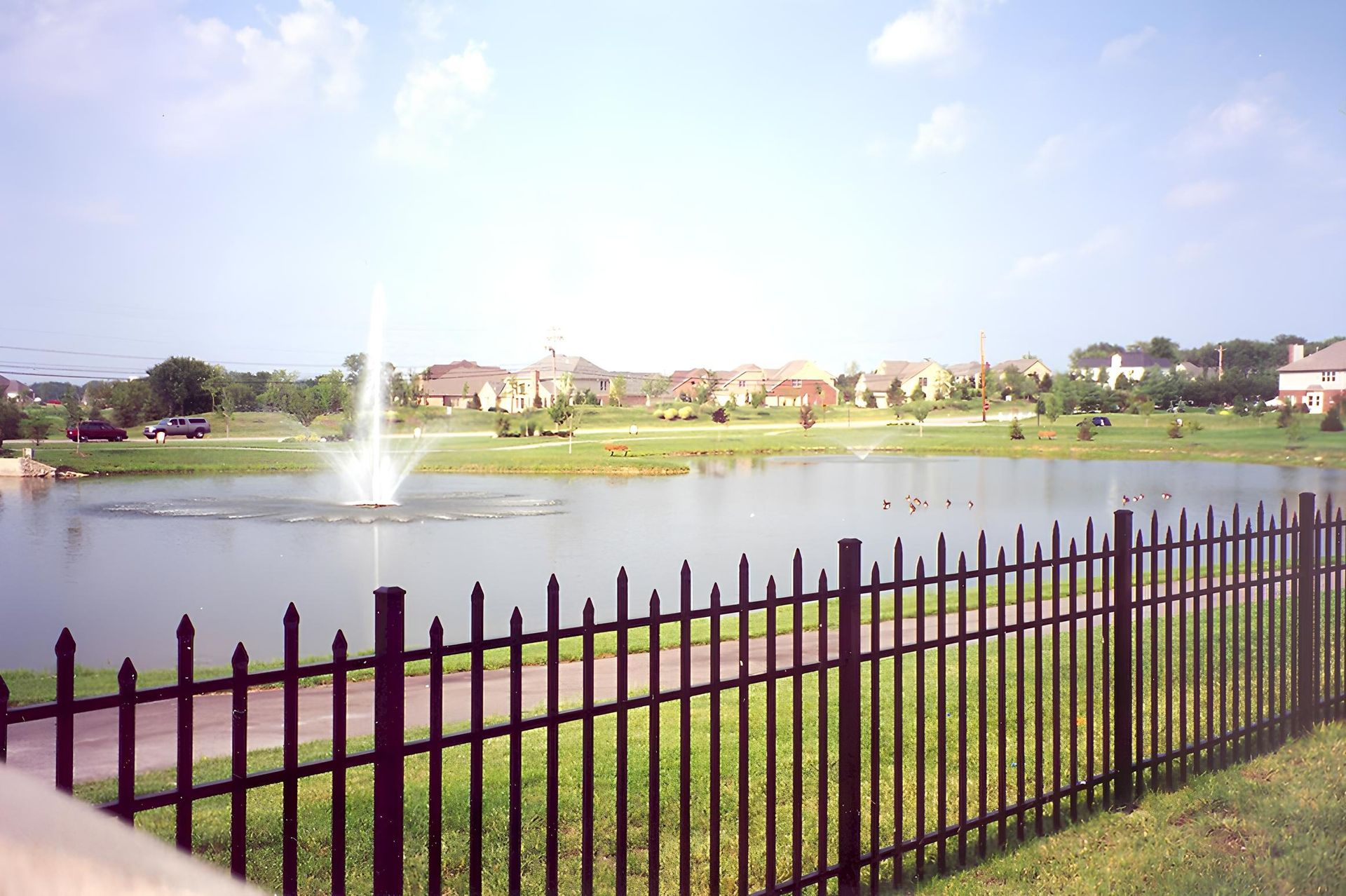 A lake with a fountain in front of a residential neighborhood; a black fence in the foreground and blue sky.