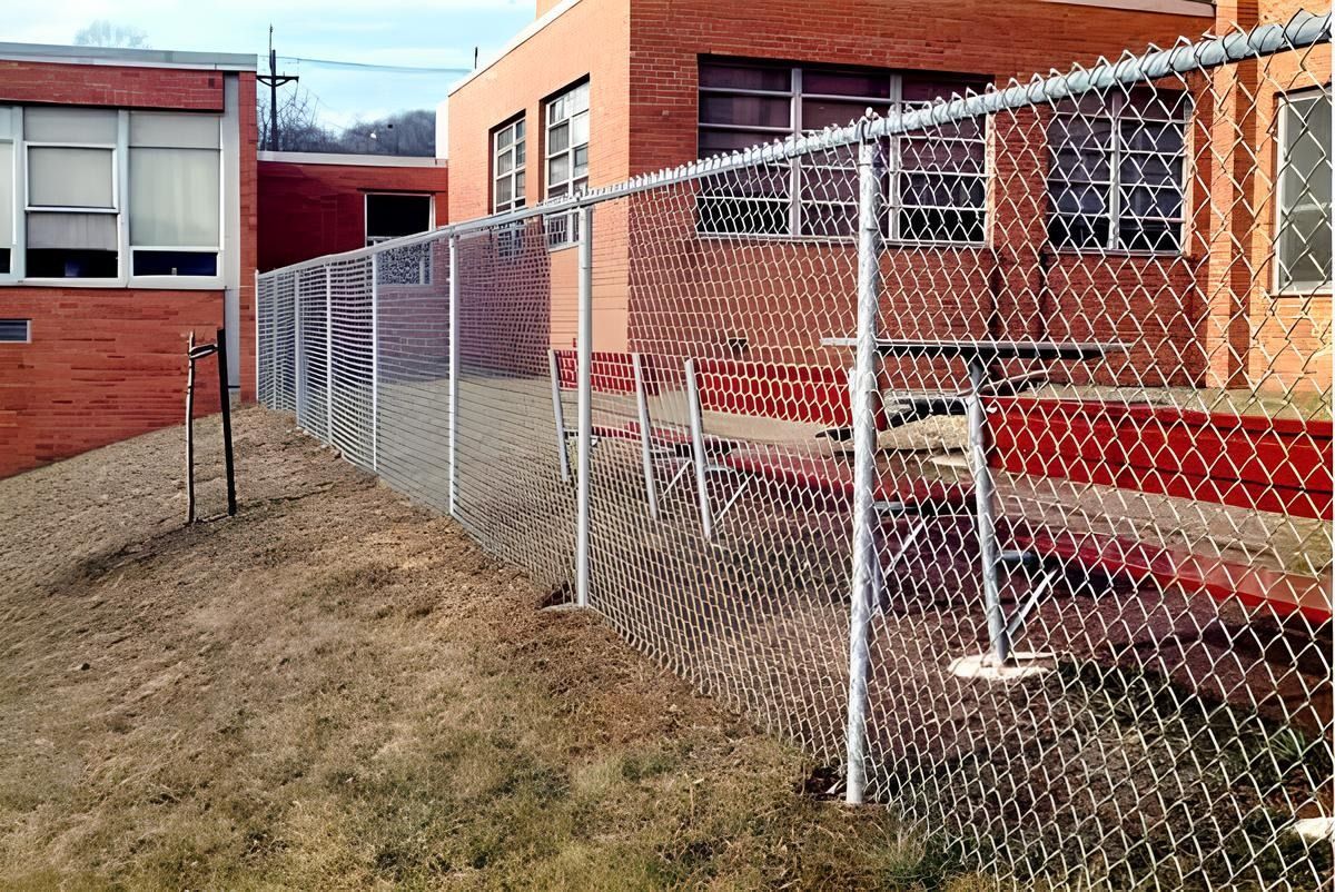 Chain-link fence along a brick building. The fence encloses a dirt area with some red seating.