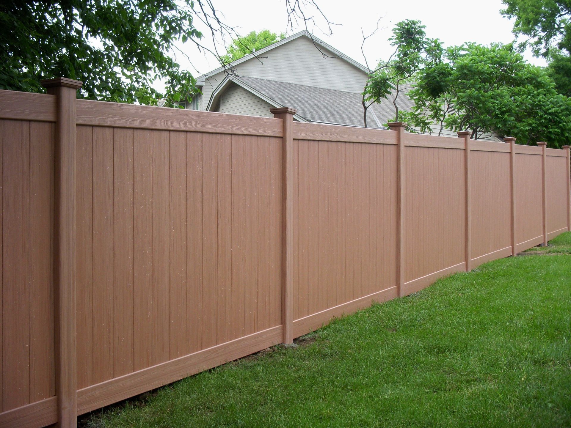 Brown vinyl privacy fence in a grassy yard, with a house and trees in the background.