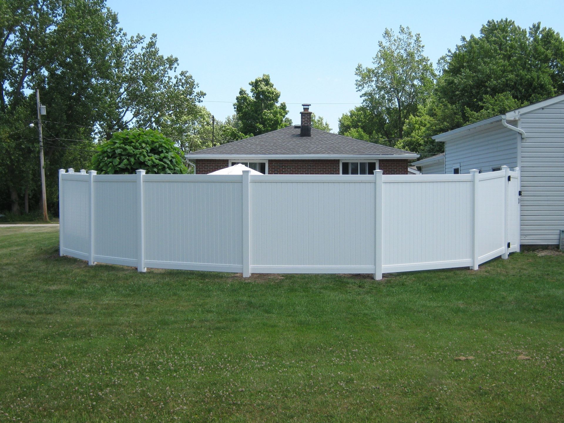 White vinyl fence enclosing a backyard, surrounding a house with a brick facade, on a sunny day.