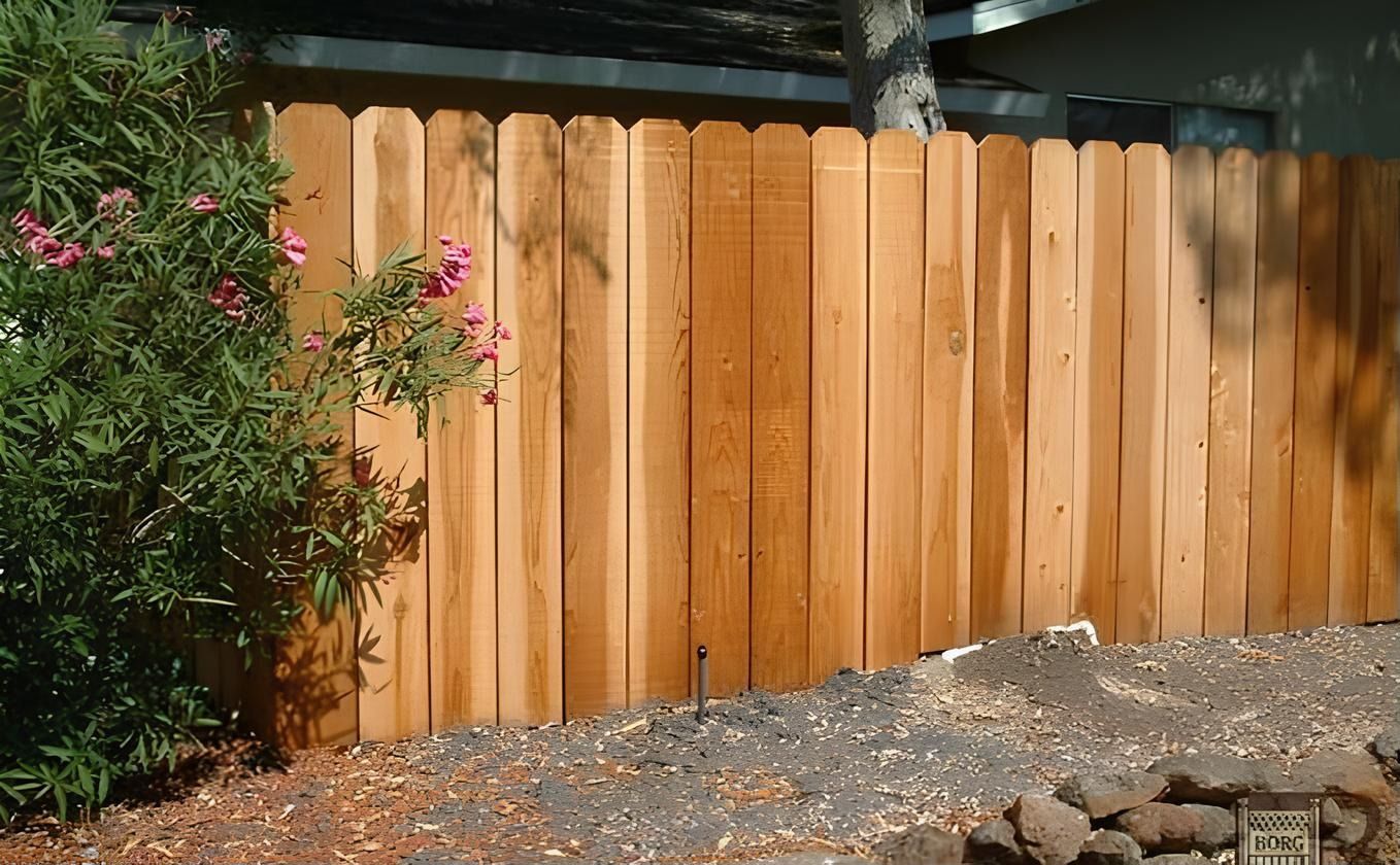 Wooden fence with vertical planks; a green bush and pink flowers are on the left.