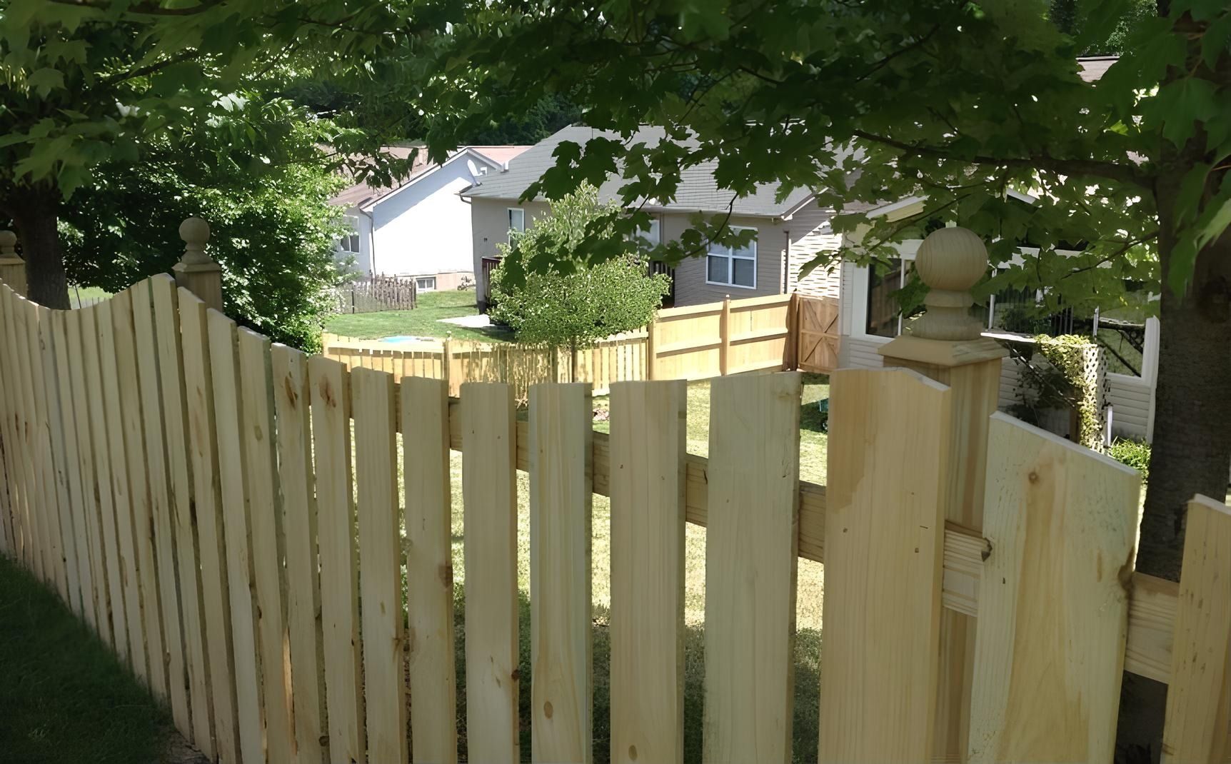 Wooden picket fence in front of a grassy yard with houses visible in the background under a tree.