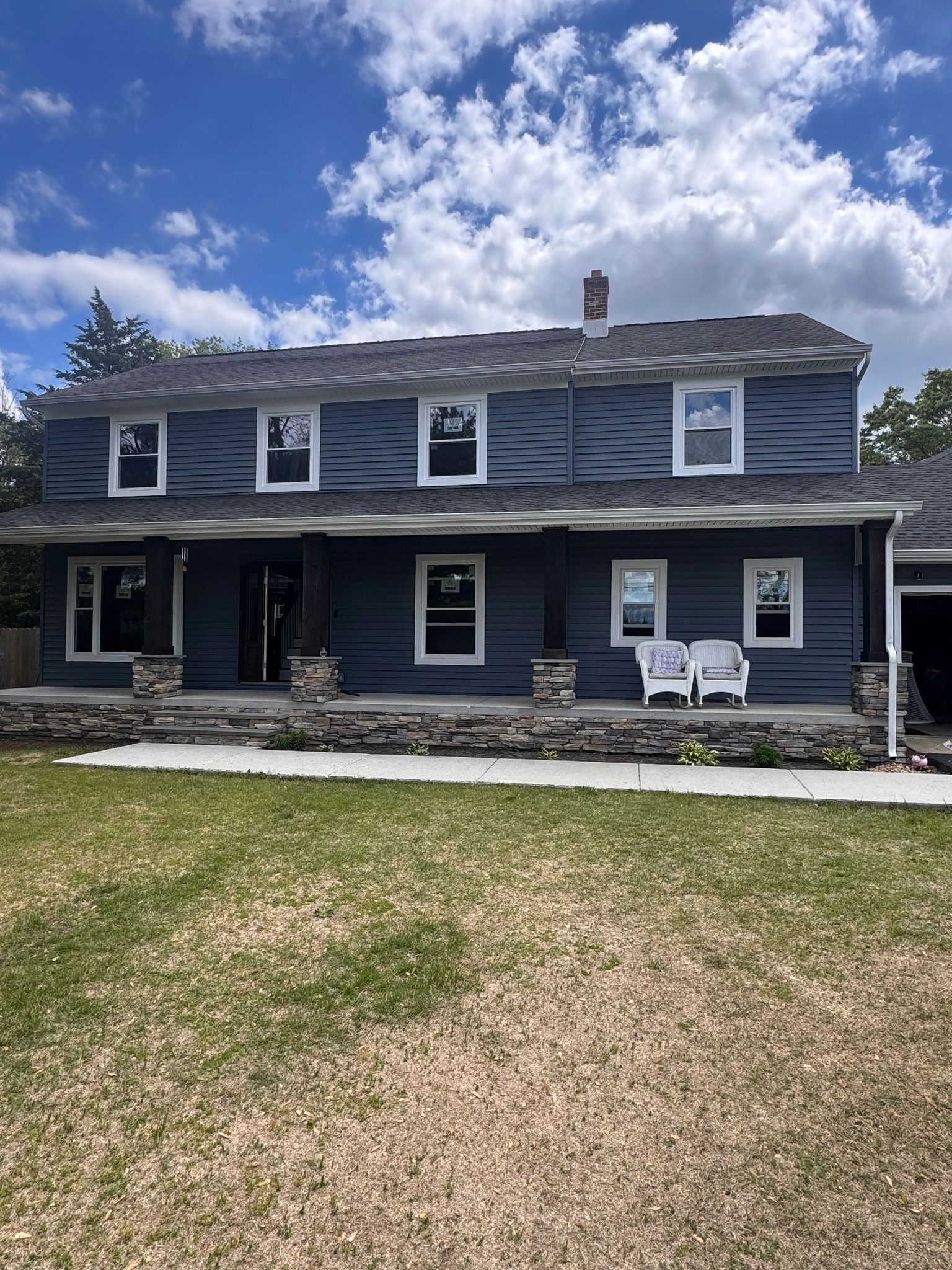 Two-story blue house with white trim. Stone porch columns, green lawn, and blue sky with clouds.