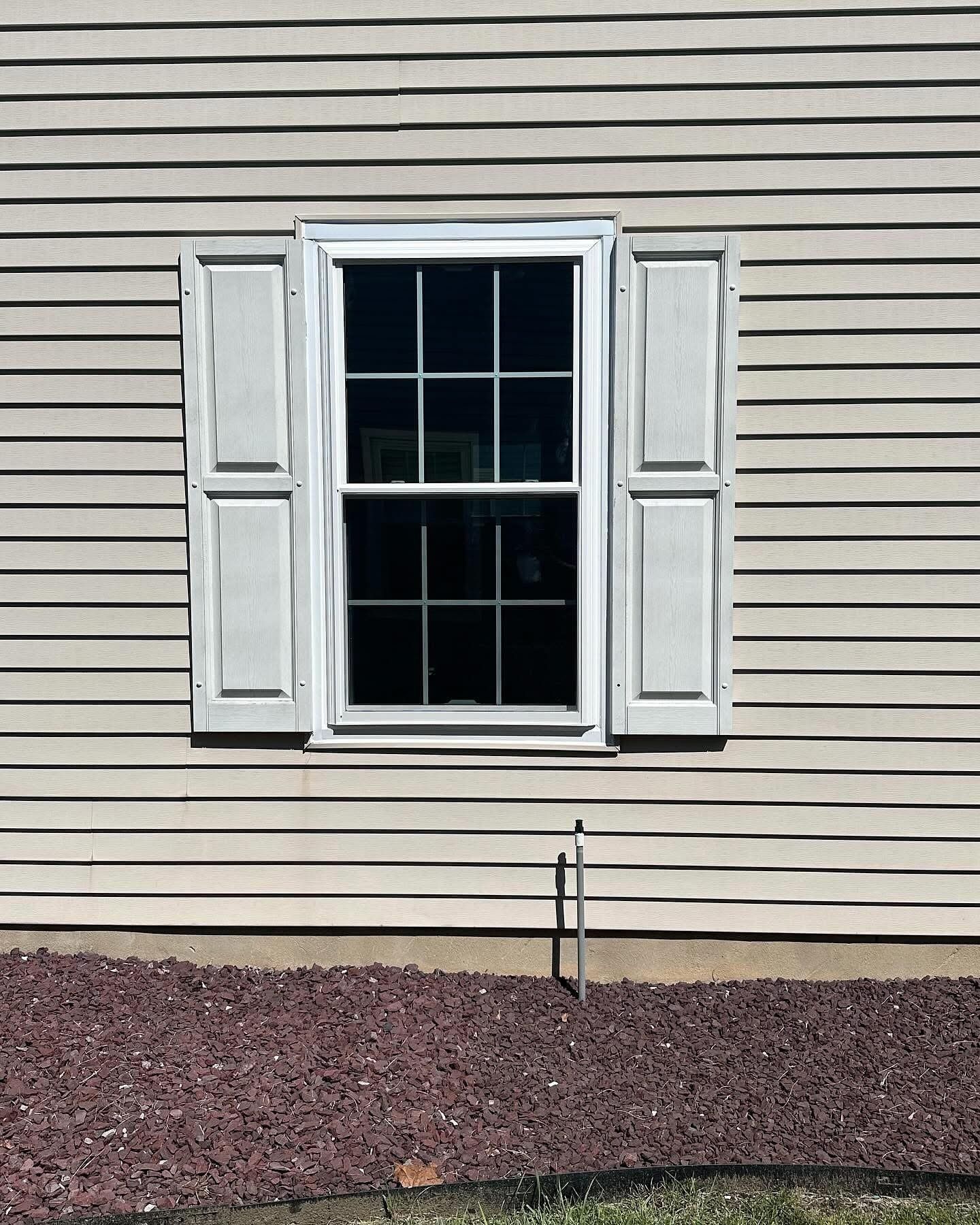 Window with white trim and shutters on beige siding, above a bed of reddish-brown stones.