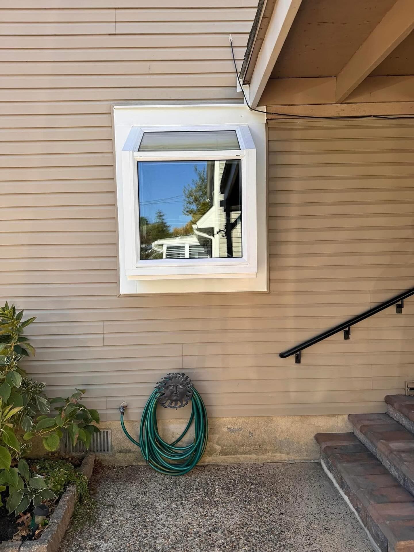 Window on a light brown building with a hose and steps. Blue sky reflected in the window.