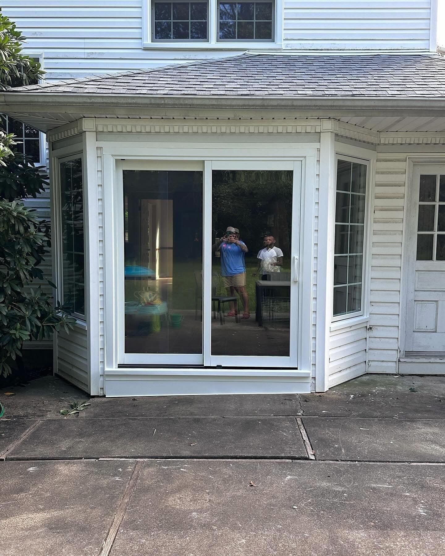 White house with a sliding glass door; two people visible in the reflection, standing outside. Concrete patio below.