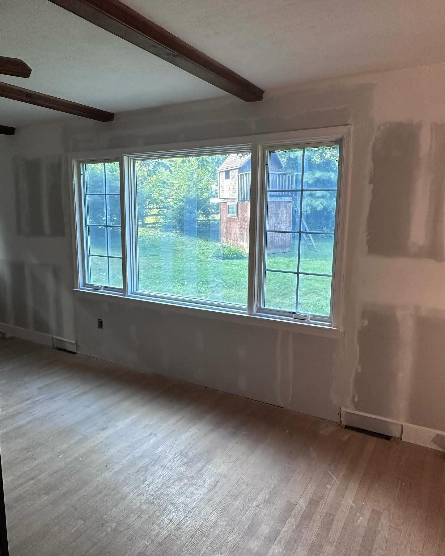 Room with large windows, hardwood floors, exposed ceiling beams, and unfinished drywall.