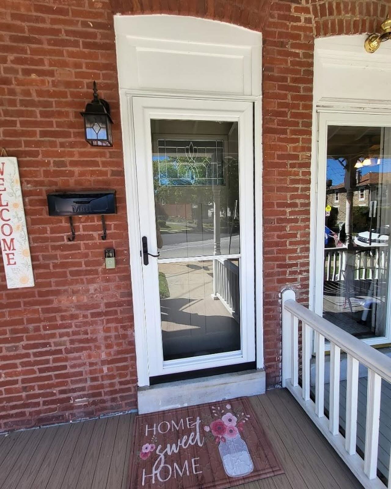 Front door with screen, red brick wall, welcome sign, and decorative doormat.
