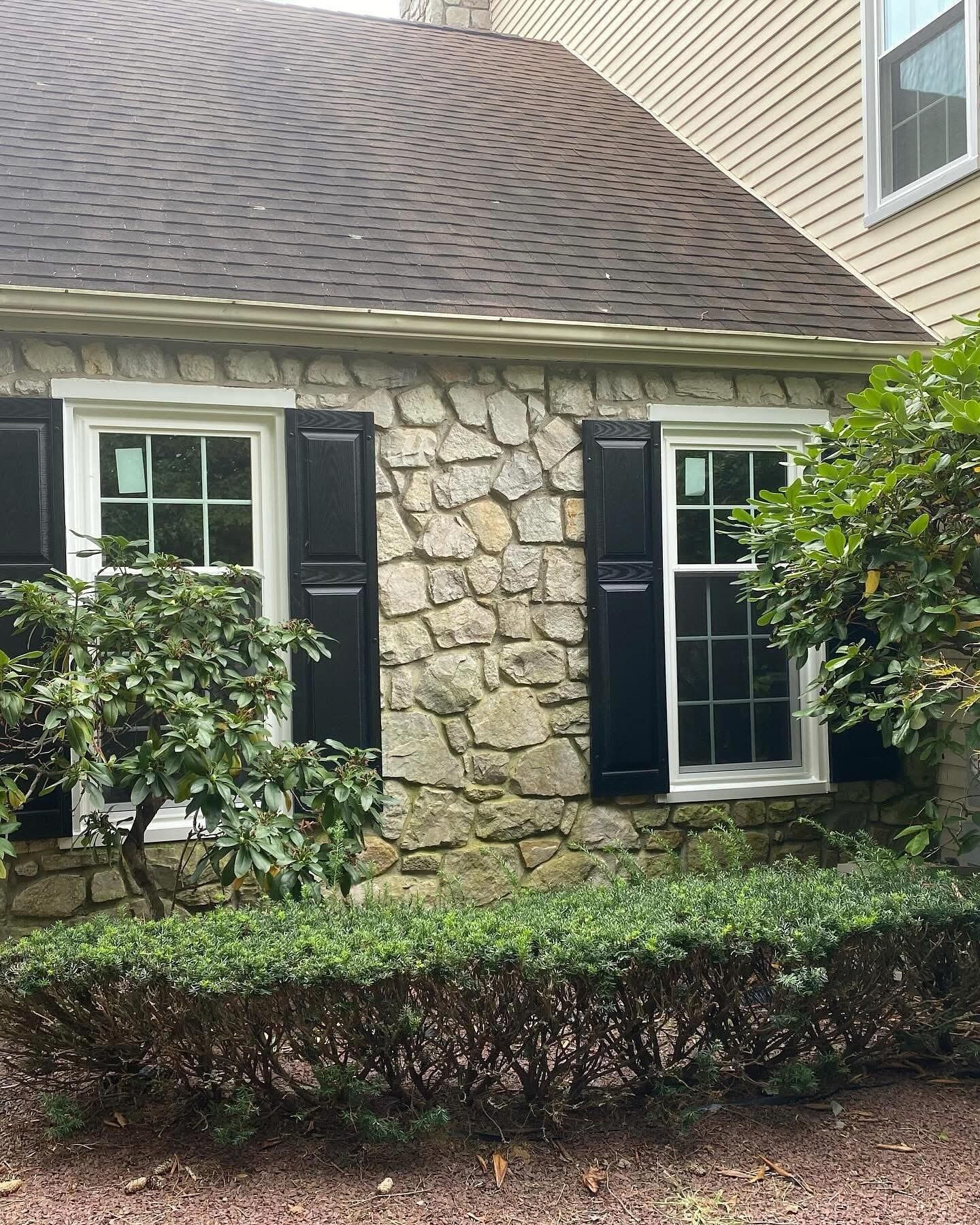 Stone house exterior with black shutters, two white-framed windows, and green shrubbery.