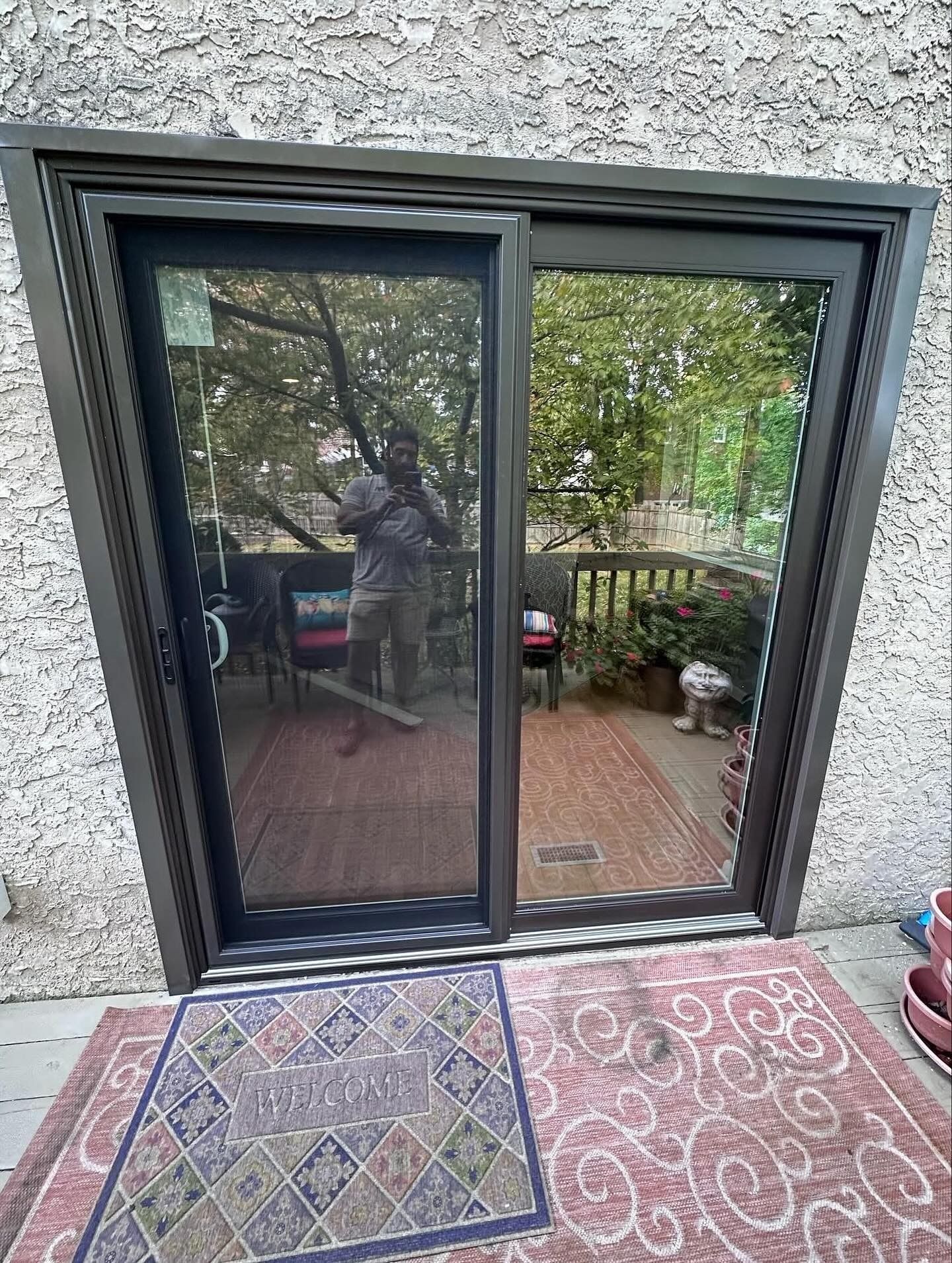 Sliding glass door, brown frame, open to a deck. A person is visible in the reflection. Red and blue rug in front.