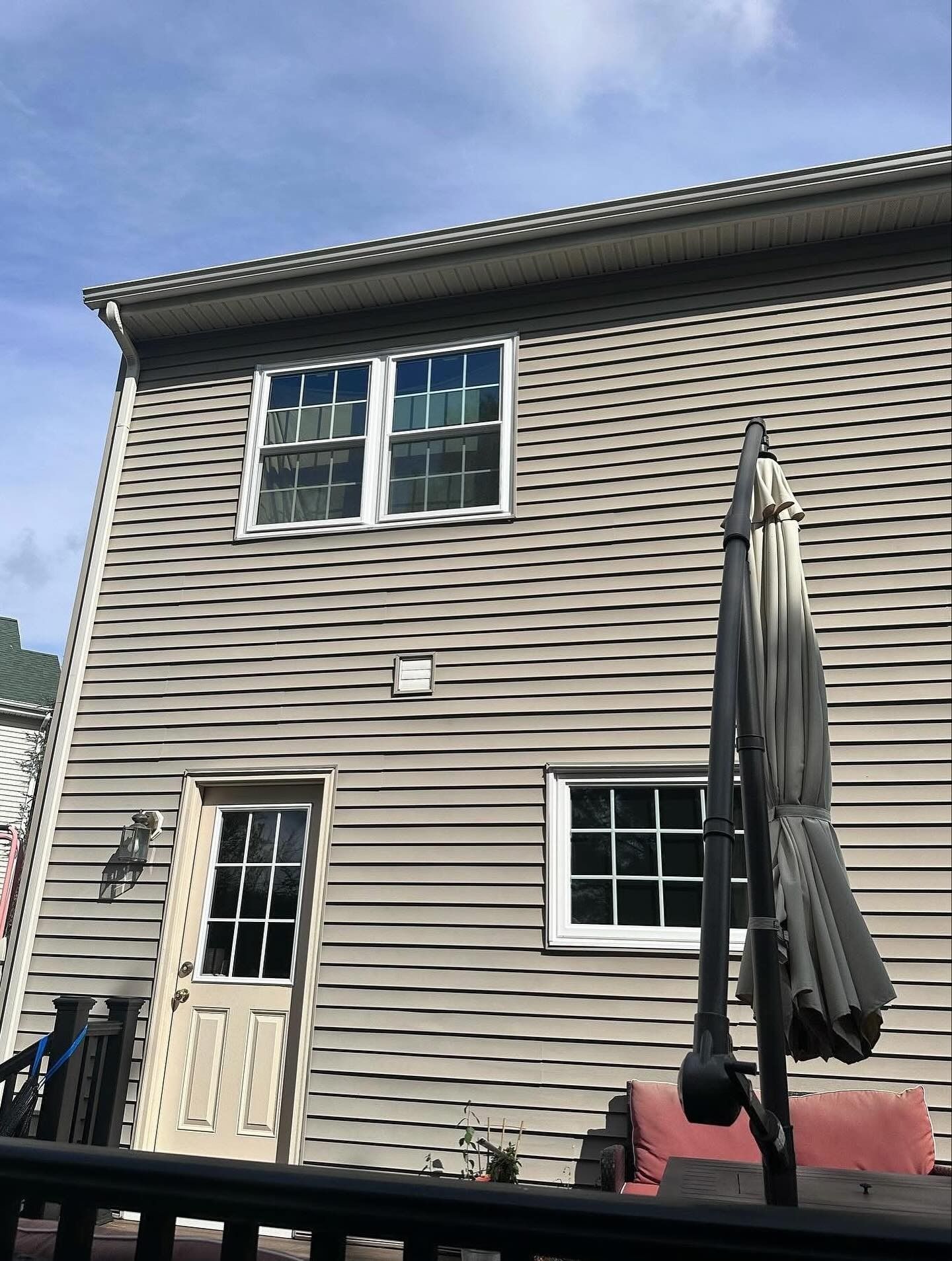 Back of a house with gray siding, windows, and a closed umbrella on a deck under a blue sky.