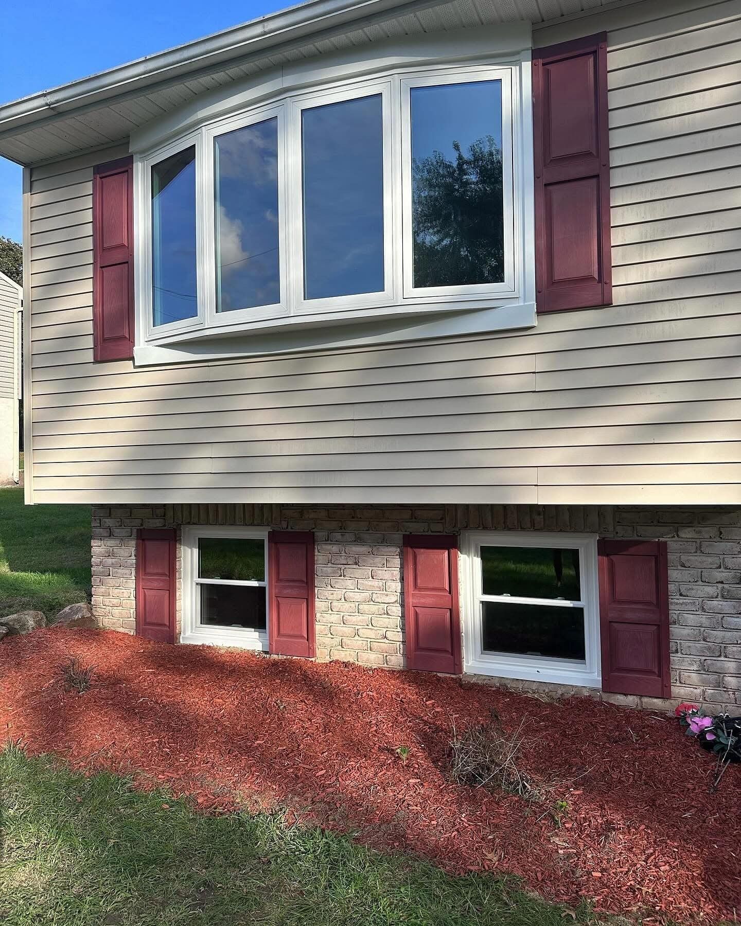 Beige house with burgundy shutters, bay window above two smaller windows. Ground covered in red mulch.