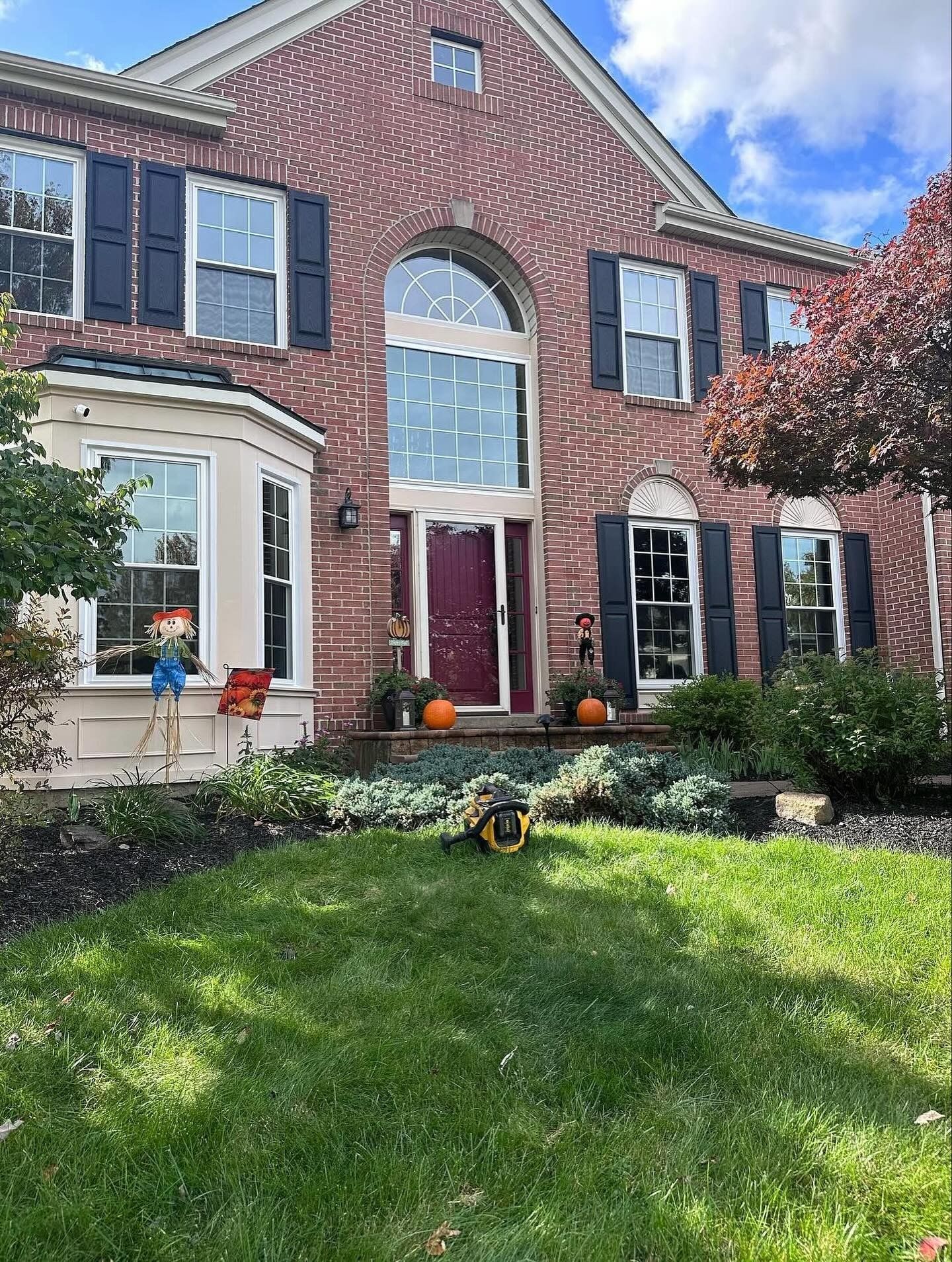 Red brick house with a dark red door and black shutters; pumpkins and a scarecrow decorate the front.