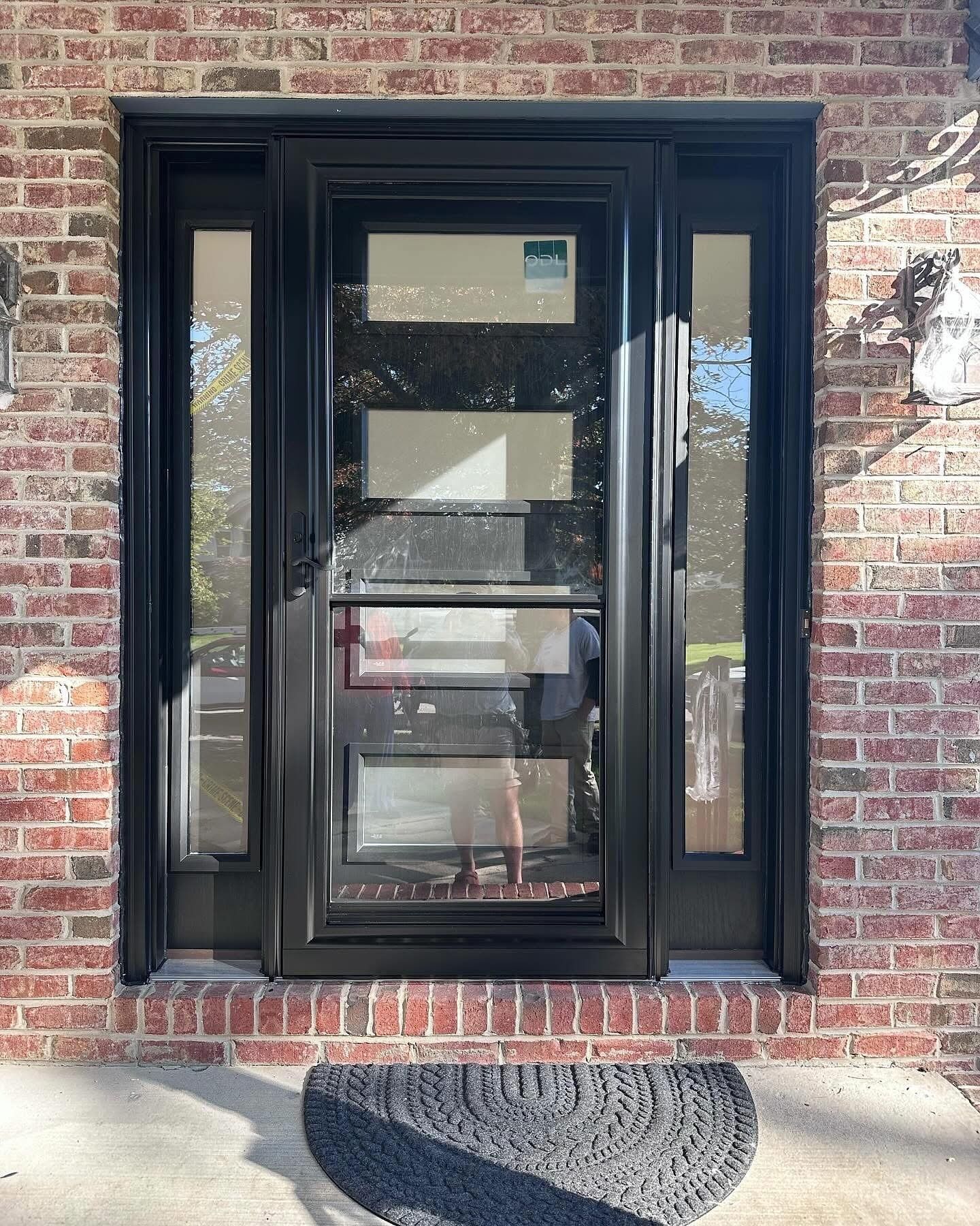 Black front door with sidelights, set in red brick. Black door mat on the concrete step.