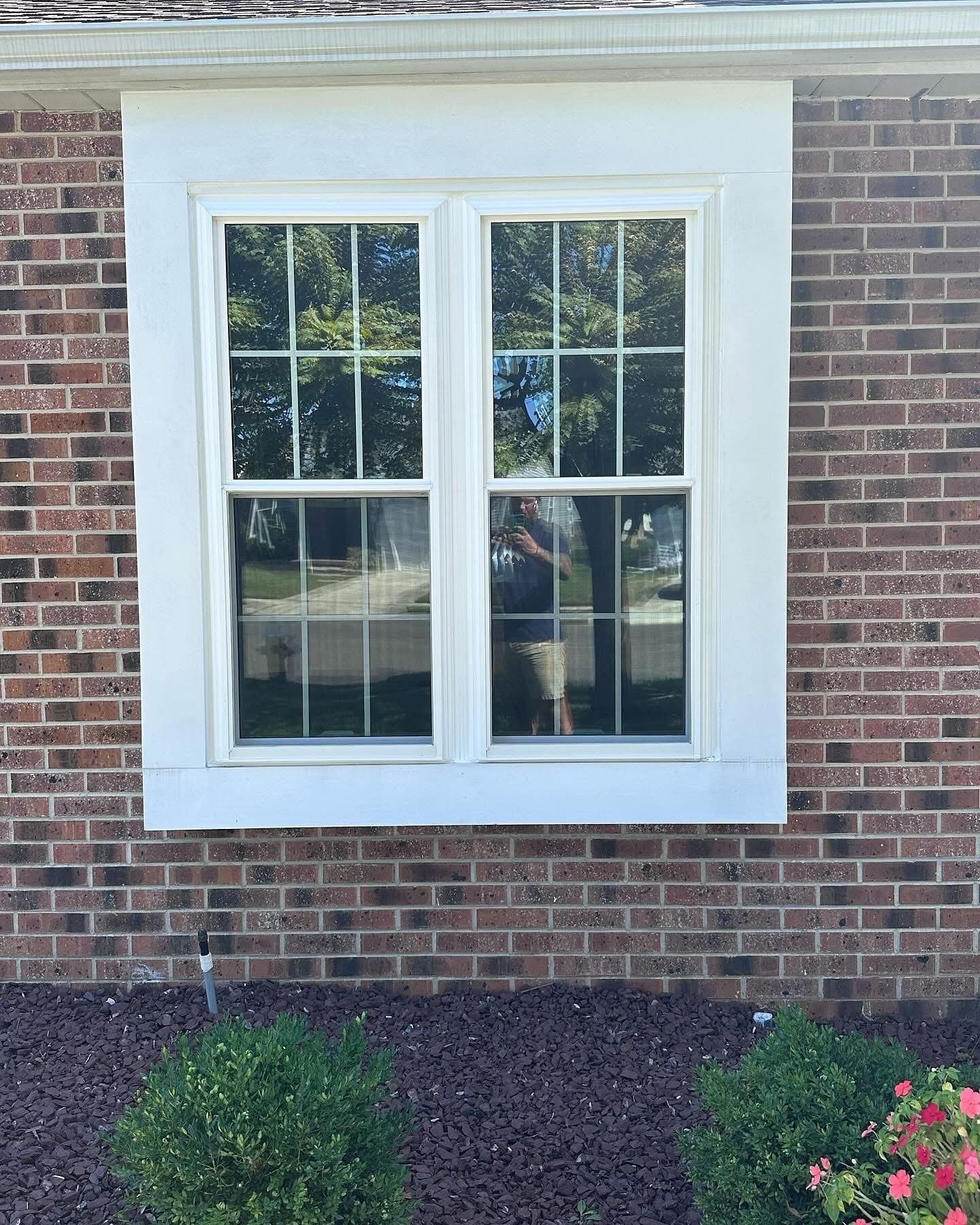 White-framed double window on a brick exterior, with the reflection of trees and a person visible.