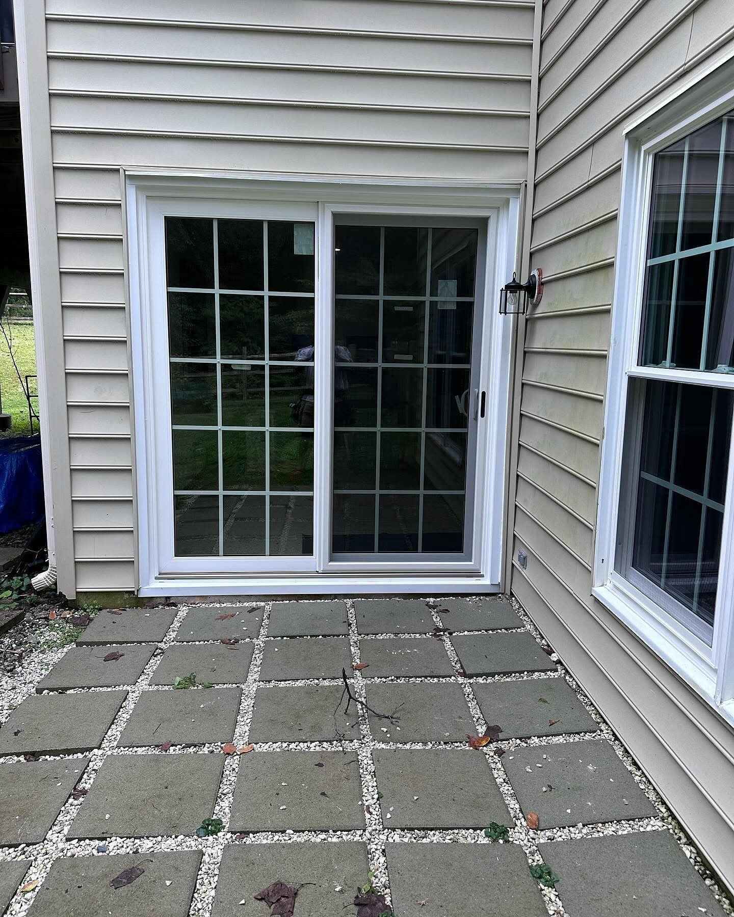 Sliding glass door with white frame on a patio with square pavers. Beige siding surrounds the door and a window.