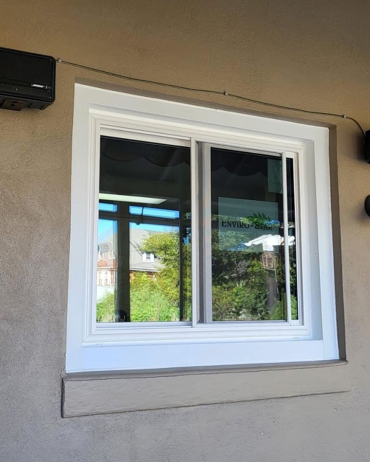 A white framed, sliding window reflecting outdoor greenery, set in a light stucco wall.