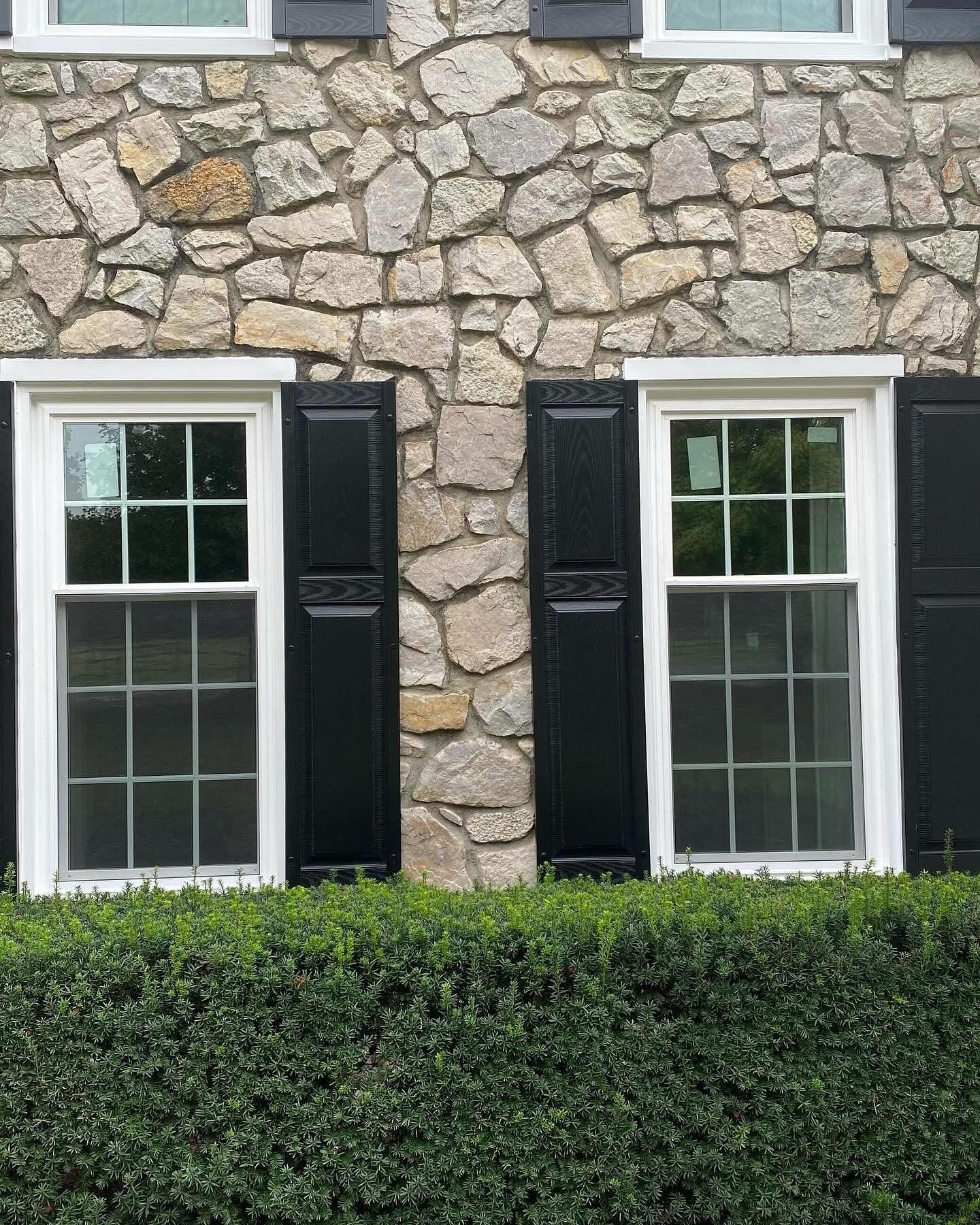 Two white-framed windows with black shutters set in a stone wall above a green hedge.