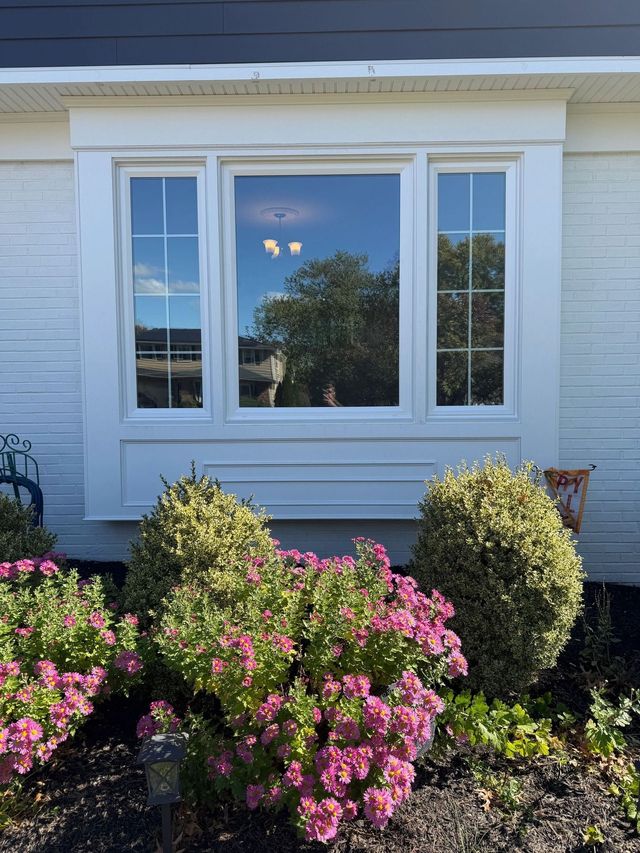 White window box with three windows reflecting sky and trees, surrounded by white trim and pink/green bushes.
