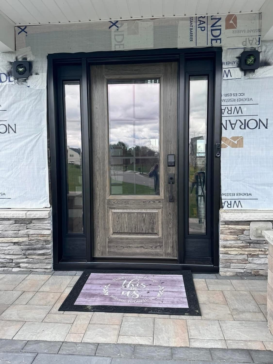 Wooden front door with sidelights, under construction, with gray and black trim and a decorative doormat.
