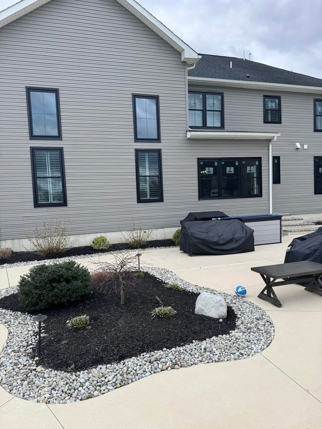 Backyard with light gray siding, dark windows, black mulch flower bed with stone border, and covered hot tub.