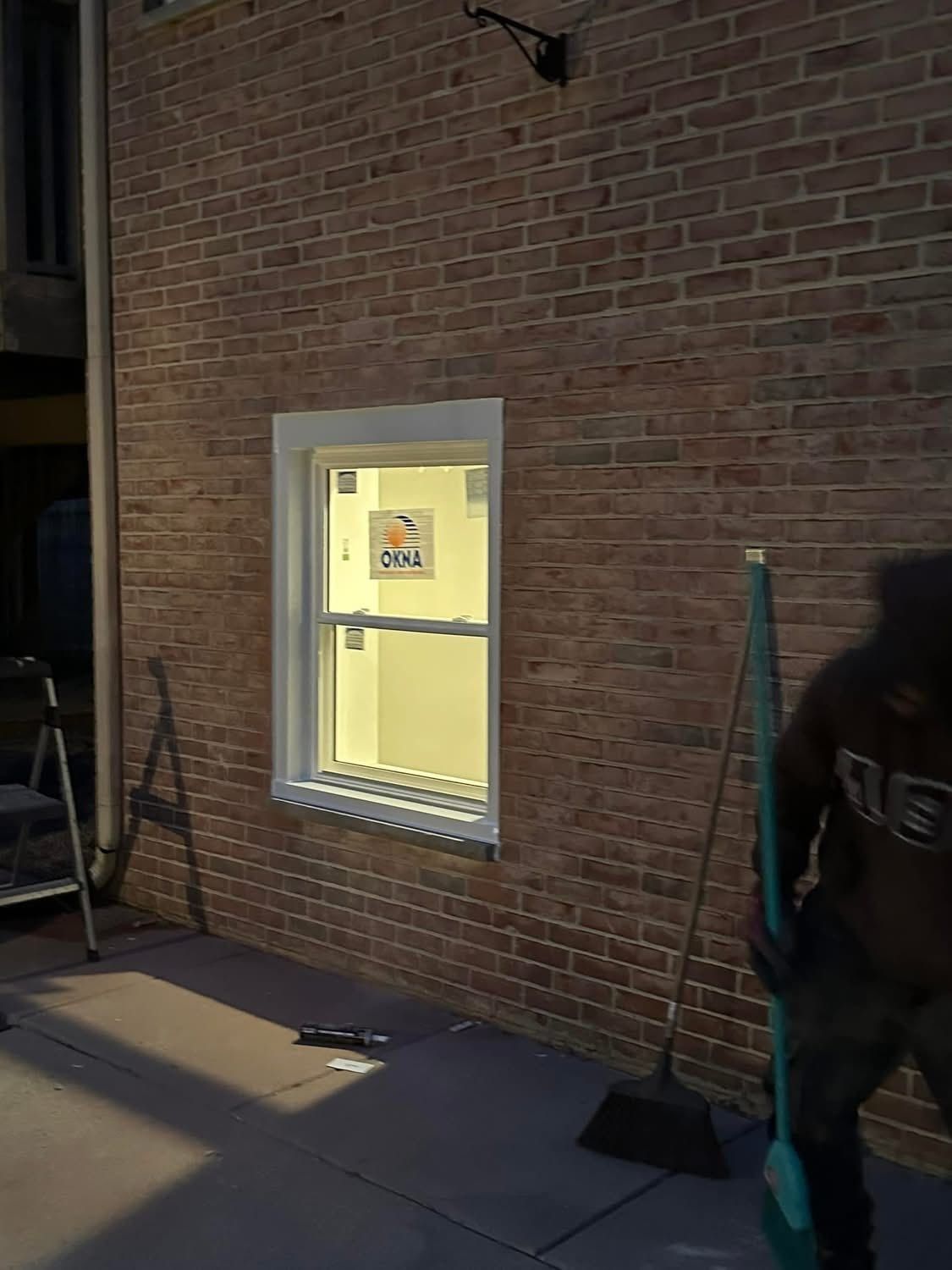 Brick building exterior with a lit window. A person with a broom stands nearby.
