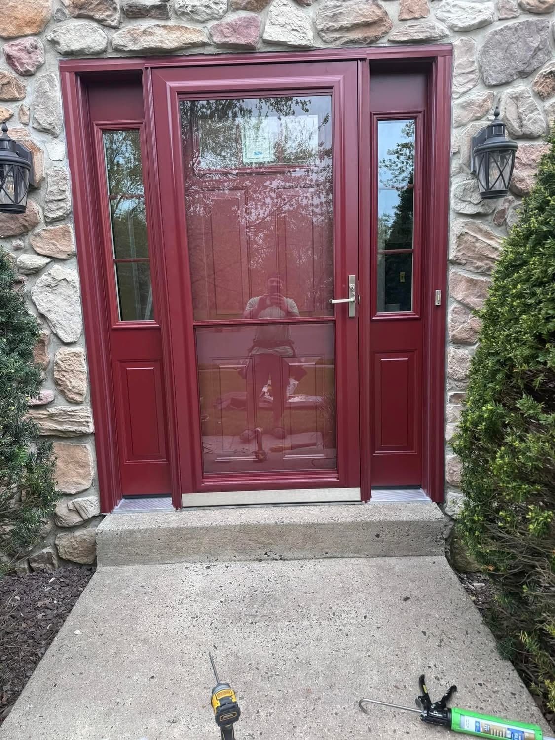 Red front door with screen, flanked by two sidelights, set in stone facade. Concrete steps lead to entrance.