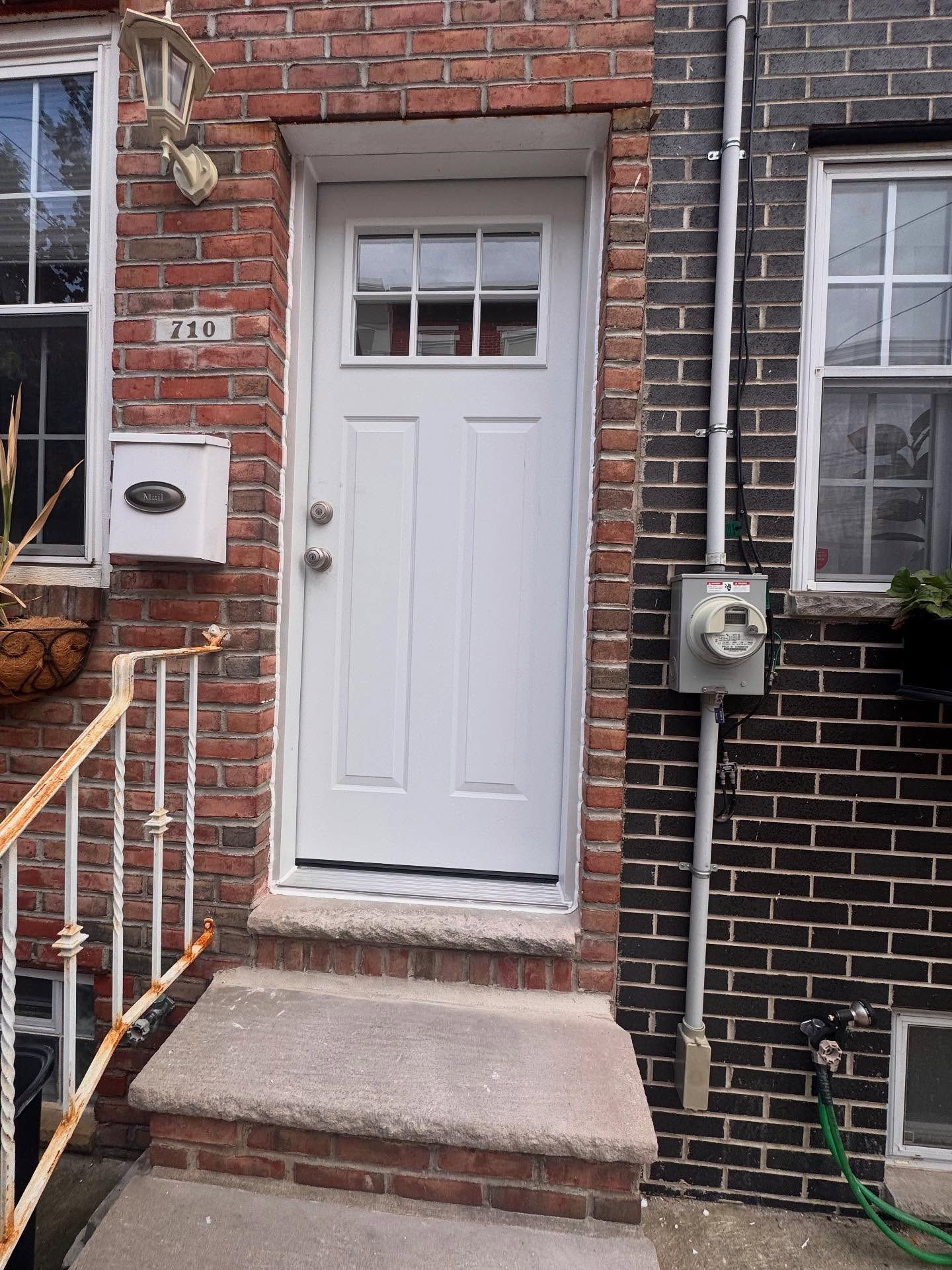 White front door with glass panel, brick and dark brick buildings, mail box, and concrete steps.