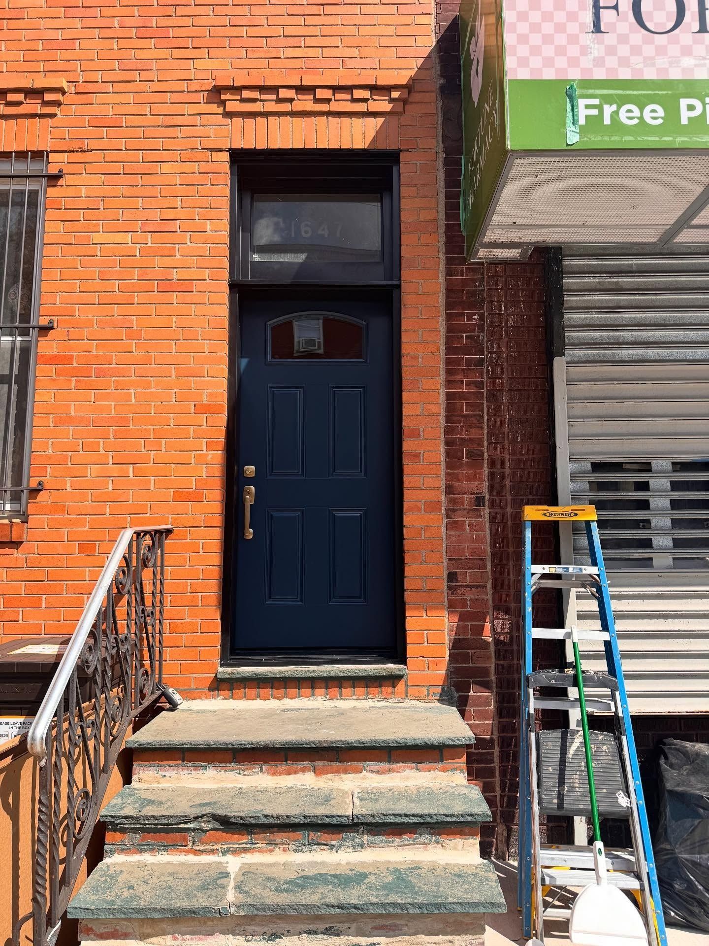 Brick building with a blue door and steps, a ladder to the side, and a metal security gate.