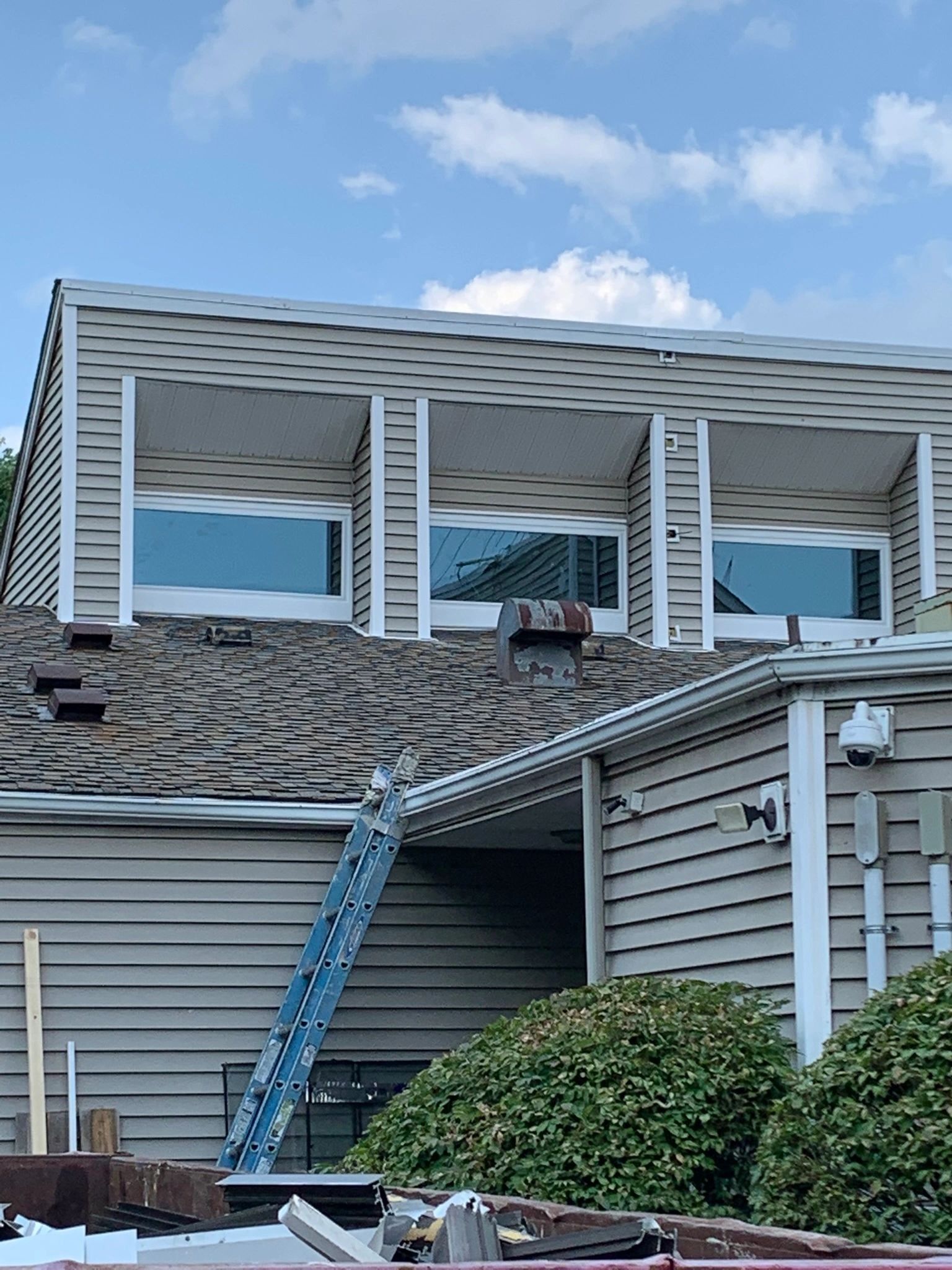 House with shingled roof, three windows, and an extending ladder. Light blue sky and green bushes visible.