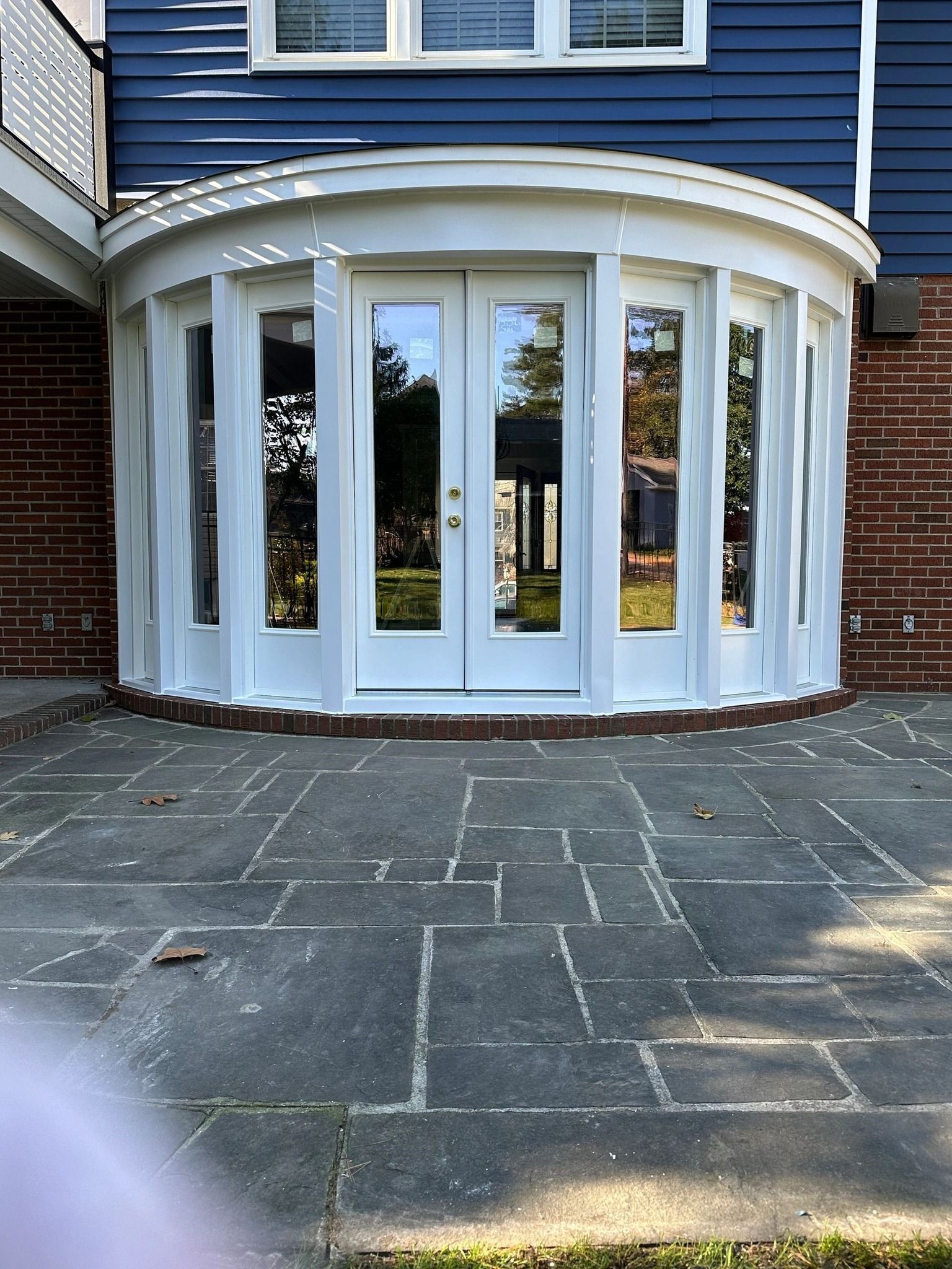 White-framed, curved sunroom with glass doors and windows, attached to a brick and blue-sided building, on a stone patio.