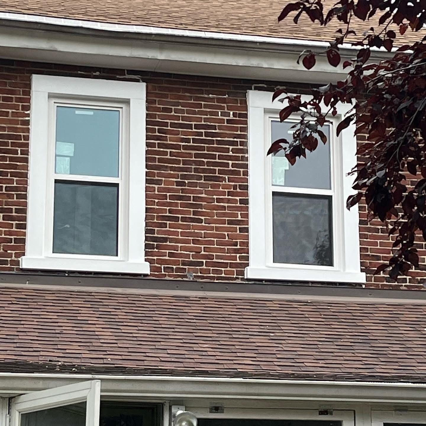Two white-framed windows on a brick building with a brown roof and burgundy tree branches.