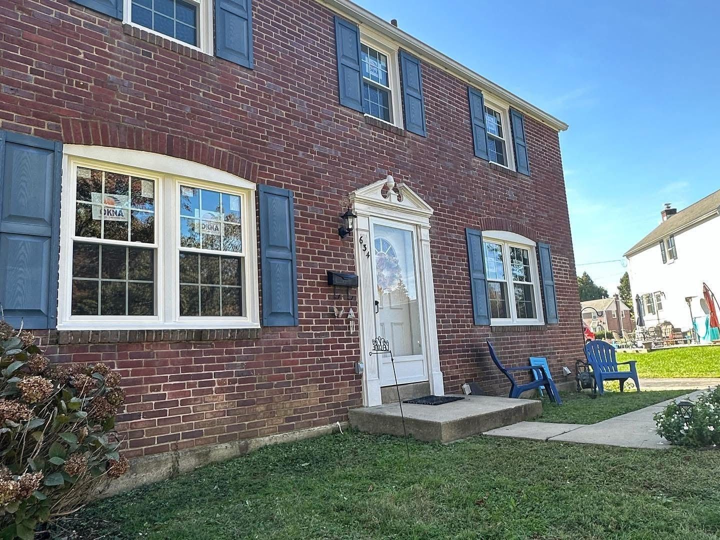 Red brick house with blue shutters, white trim, and a small porch. Two blue chairs sit on the grass.