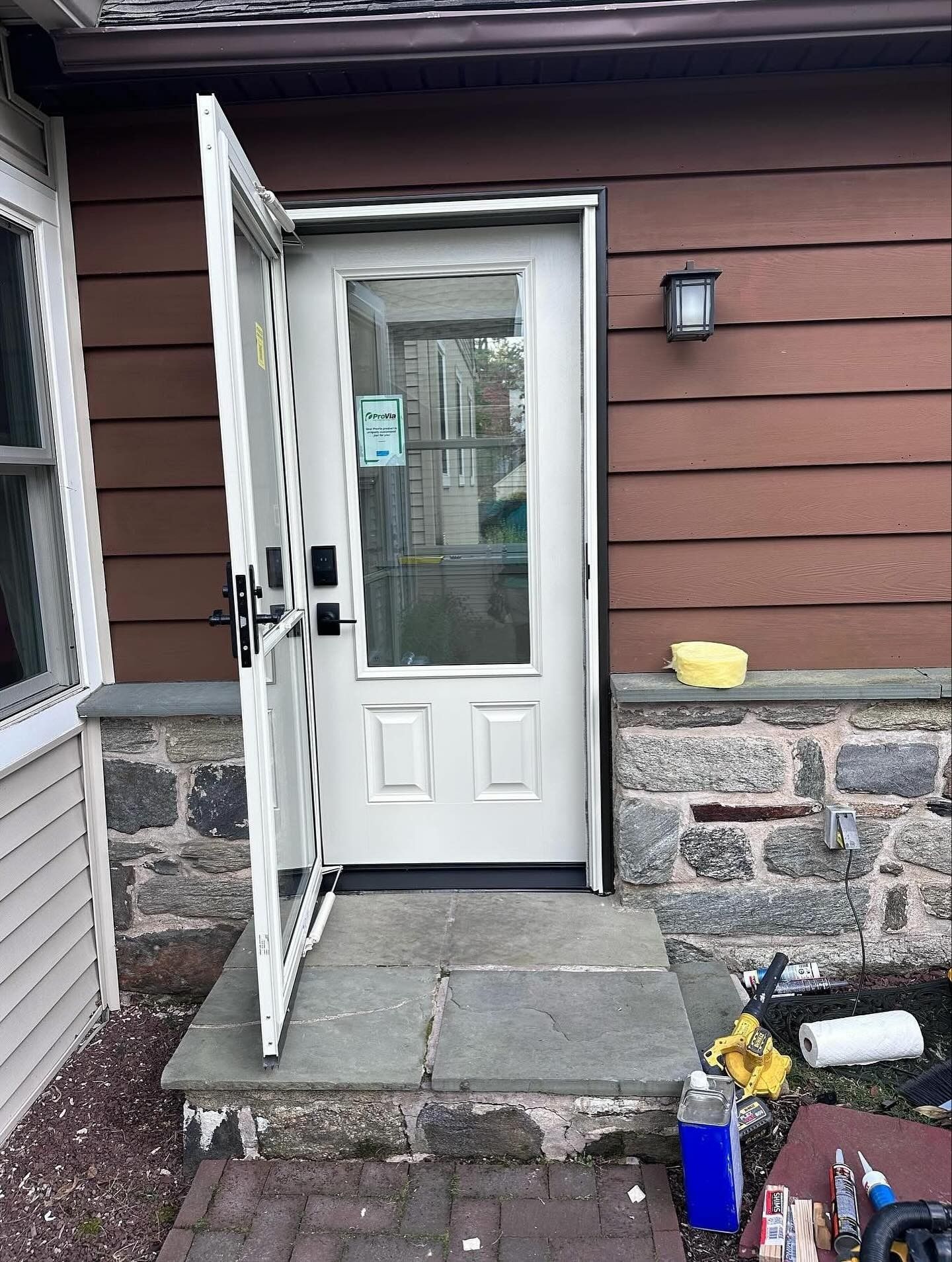 Open white door on stone steps; brown siding, brick foundation, tools nearby.