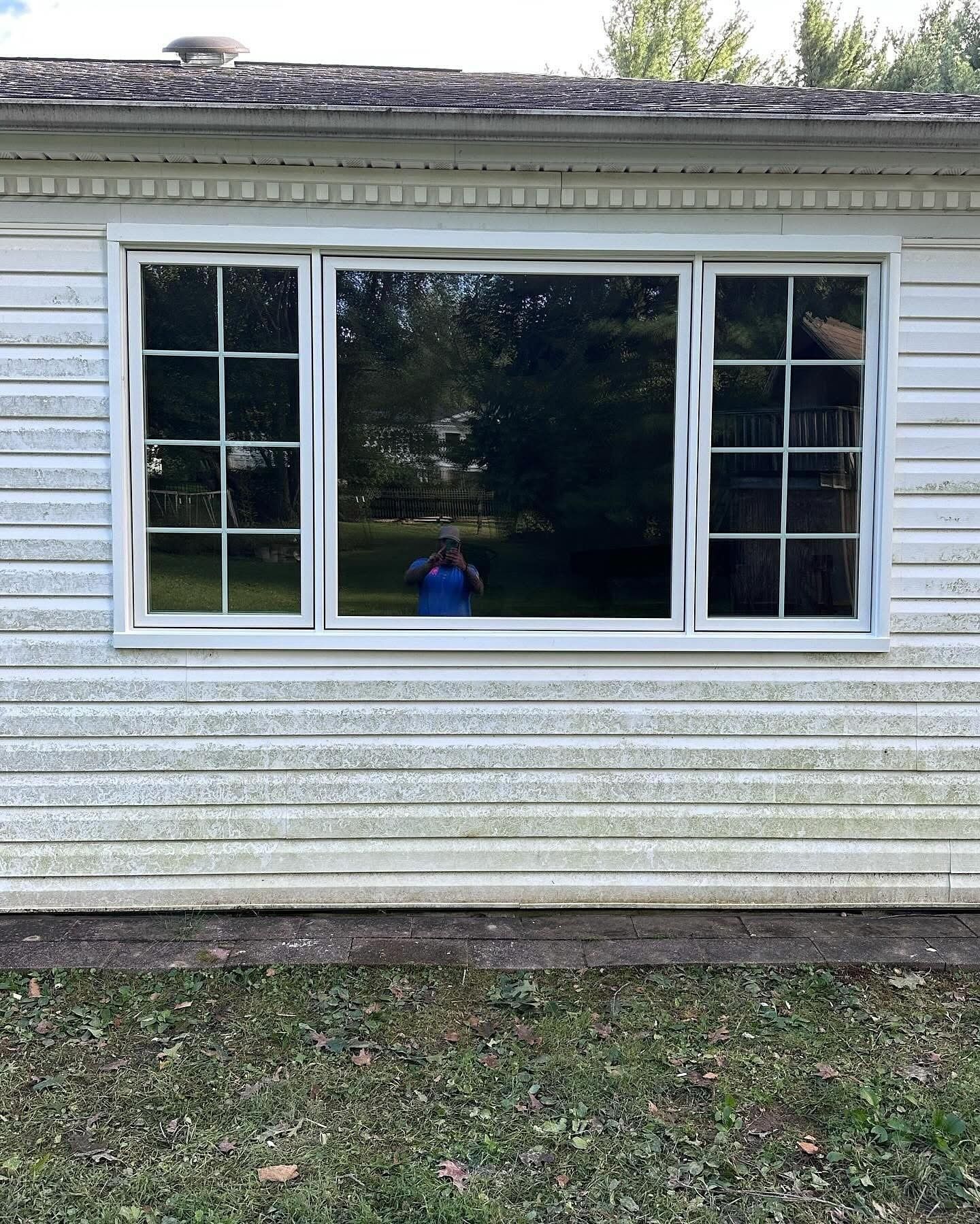 White framed window in a white-sided building; a person is reflected in the glass, outside on grass.