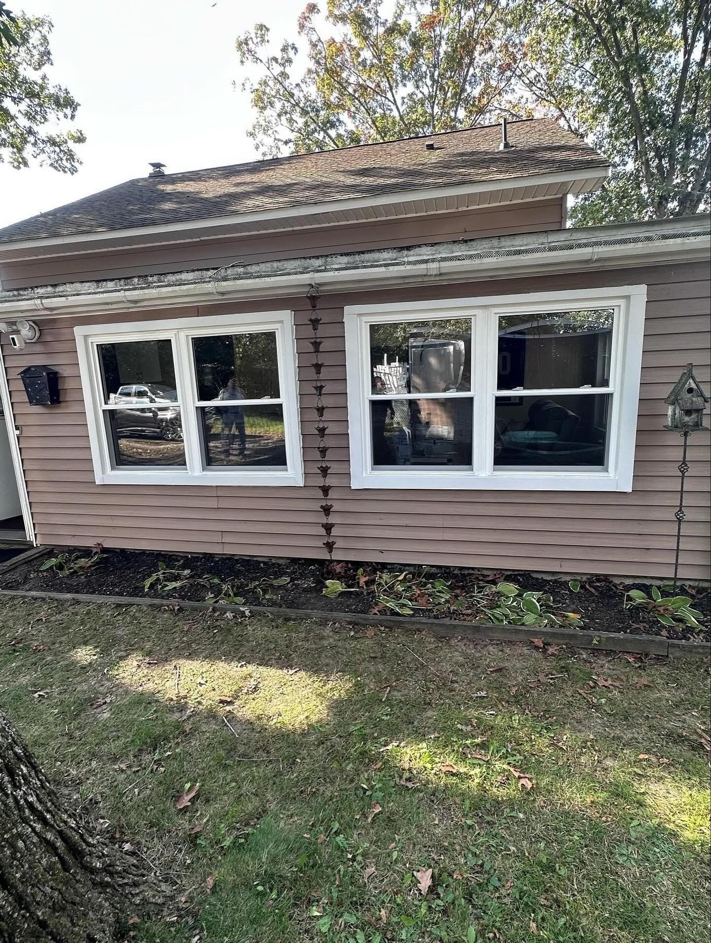 A light-brown house with two white-framed windows, surrounded by grass and a garden bed.