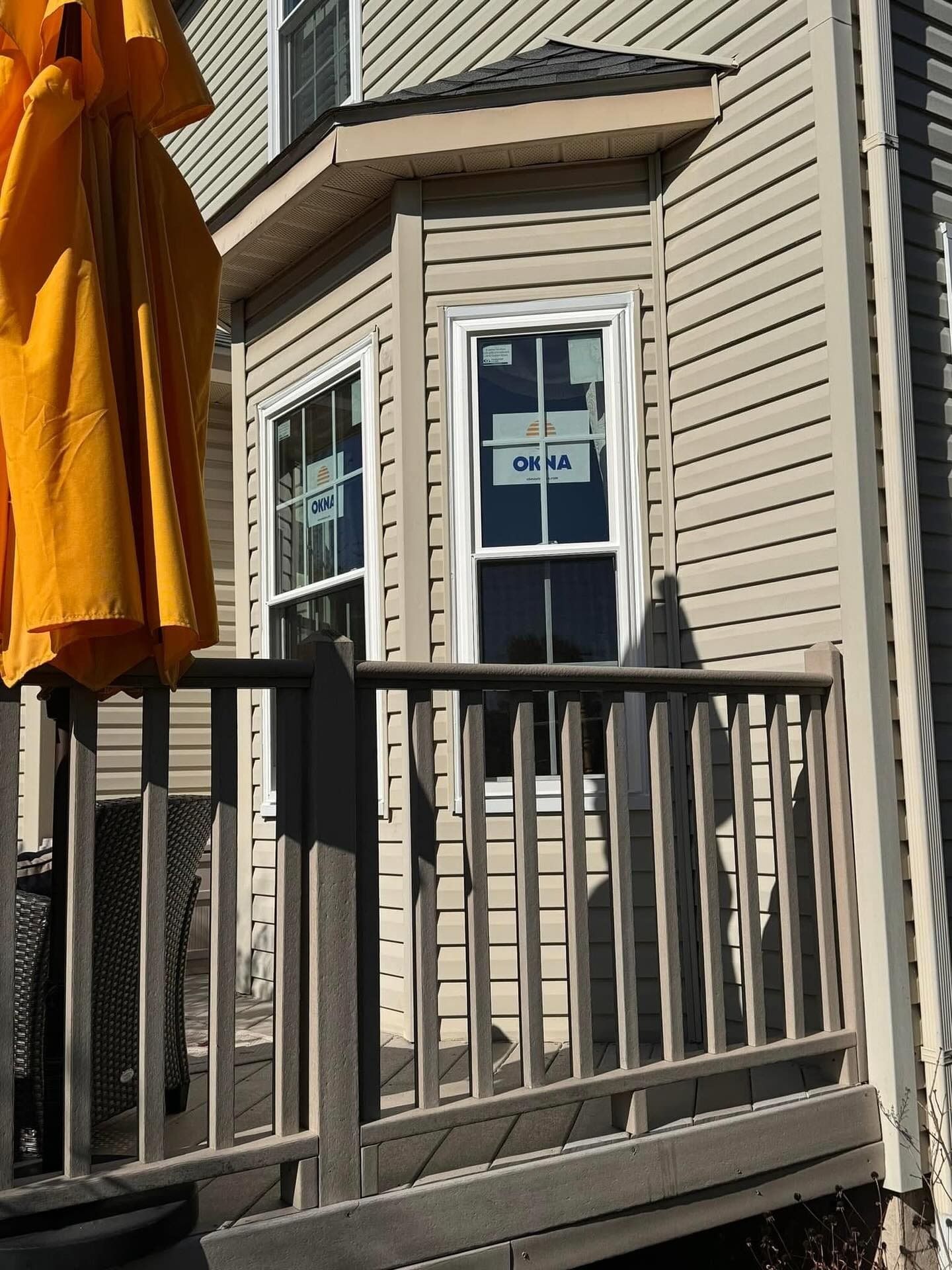 Bay window with two vertical windows in a light tan sided house.  Deck in front of the window with umbrella on left side.