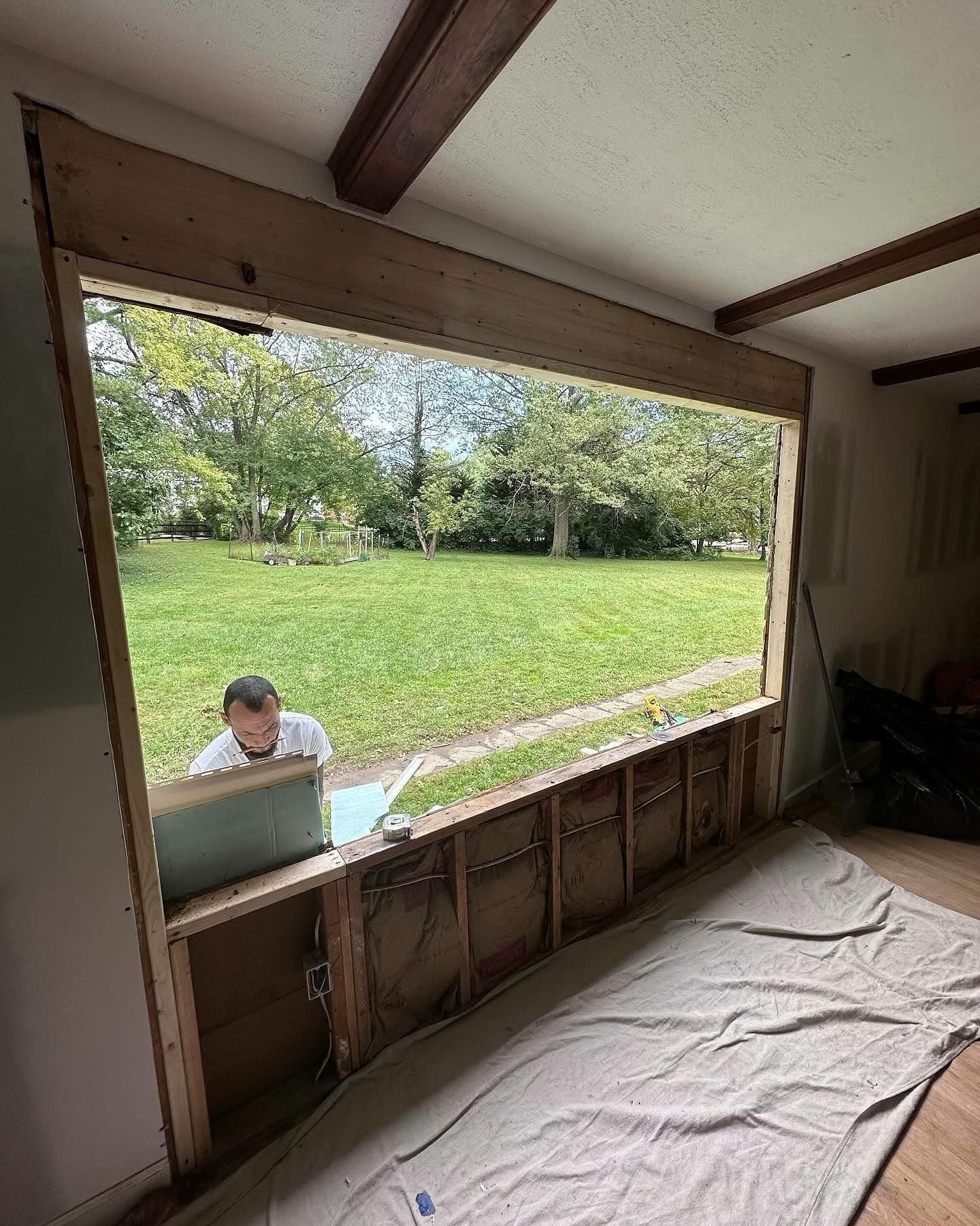 Man working on a large window frame, interior view. Green yard visible through opening.