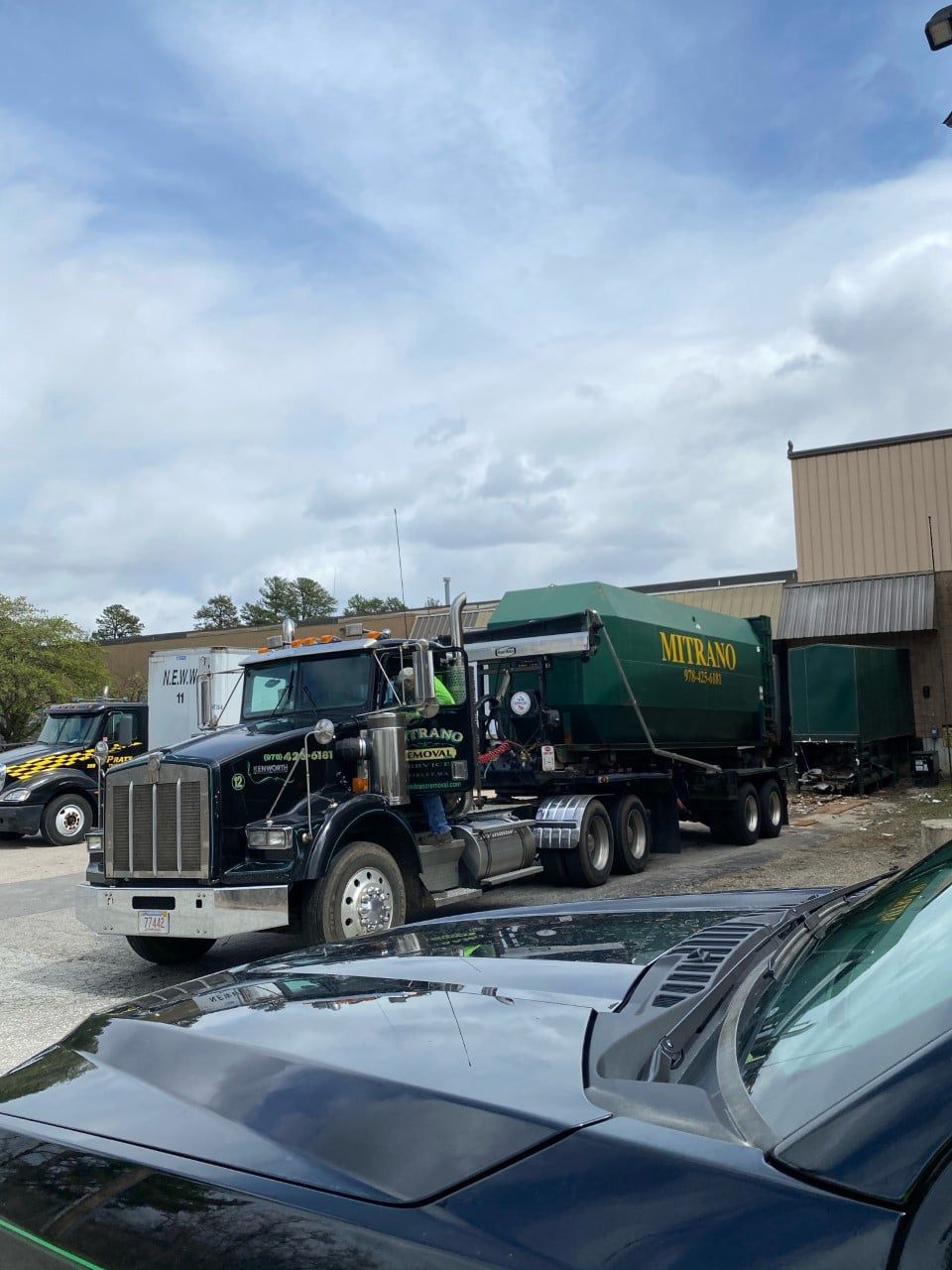 Black semi-truck hauling a green trailer; parked near a building. Overcast sky.